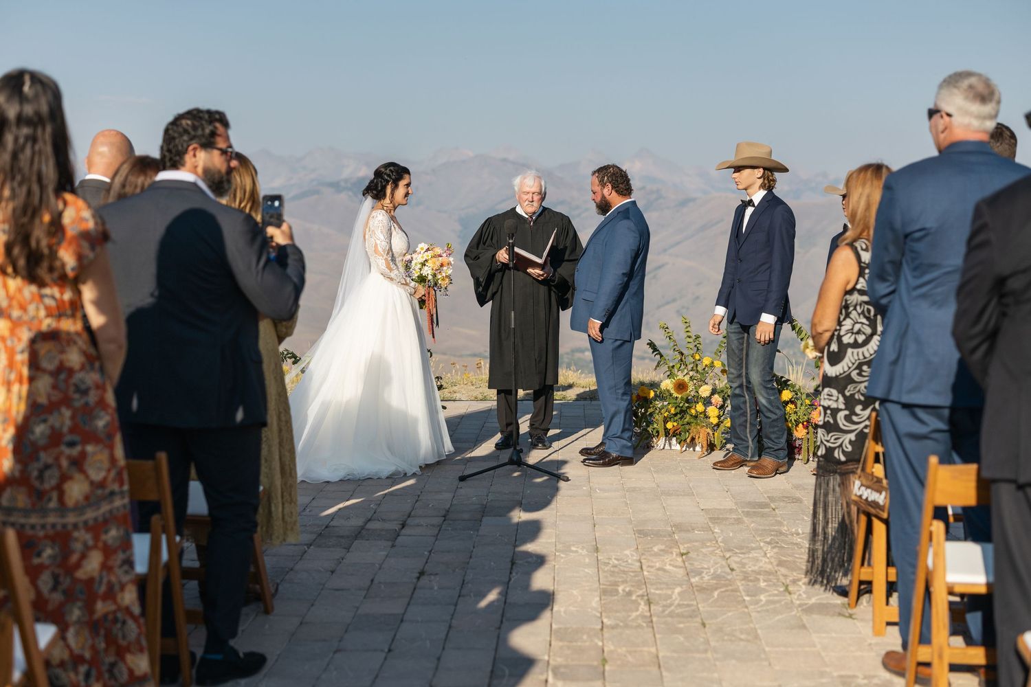 A wedding ceremony takes place on a stone patio with mountain views during a clear sunny day.