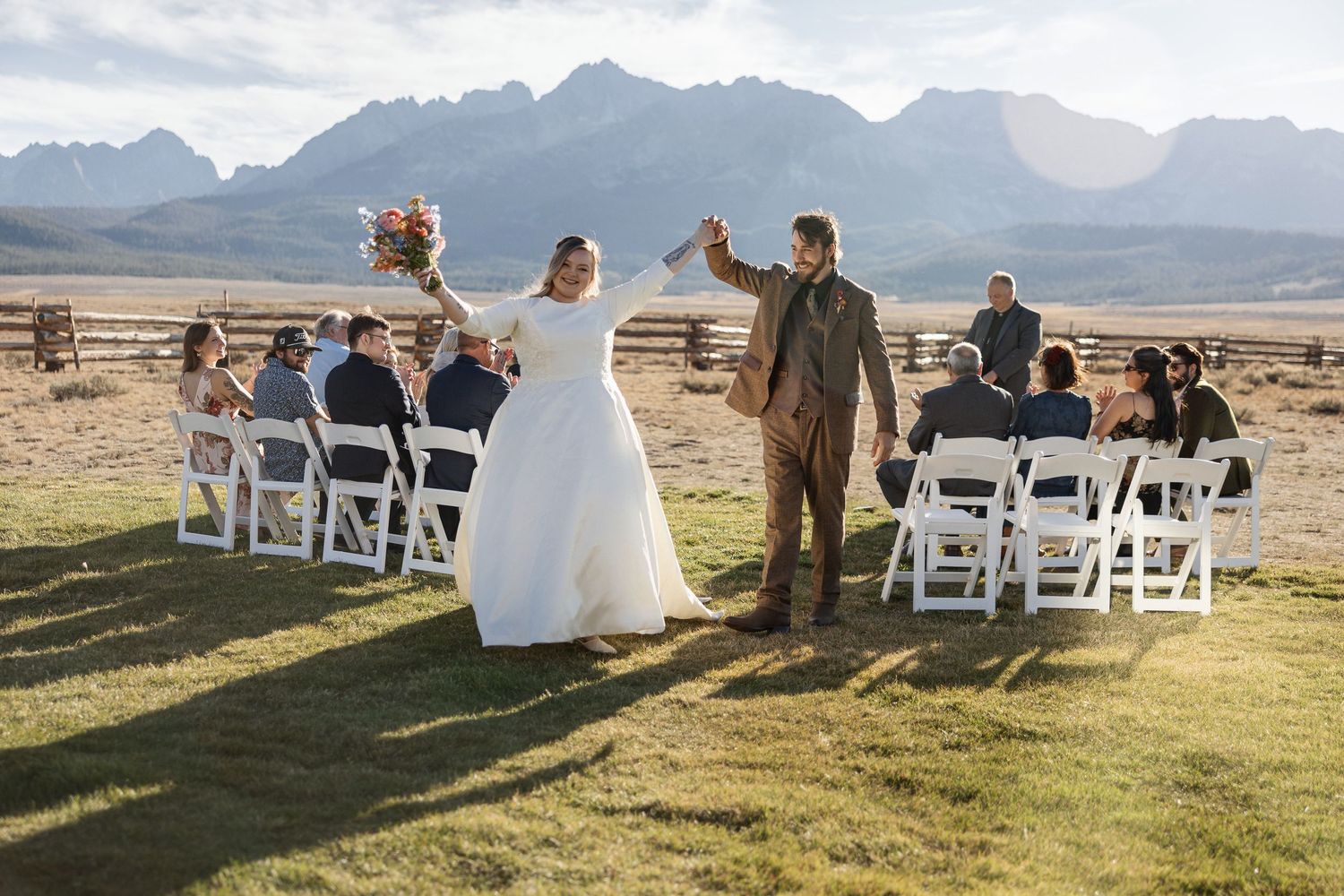 A small outdoor wedding ceremony takes place against majestic mountain scenery with white chairs set up on grass.