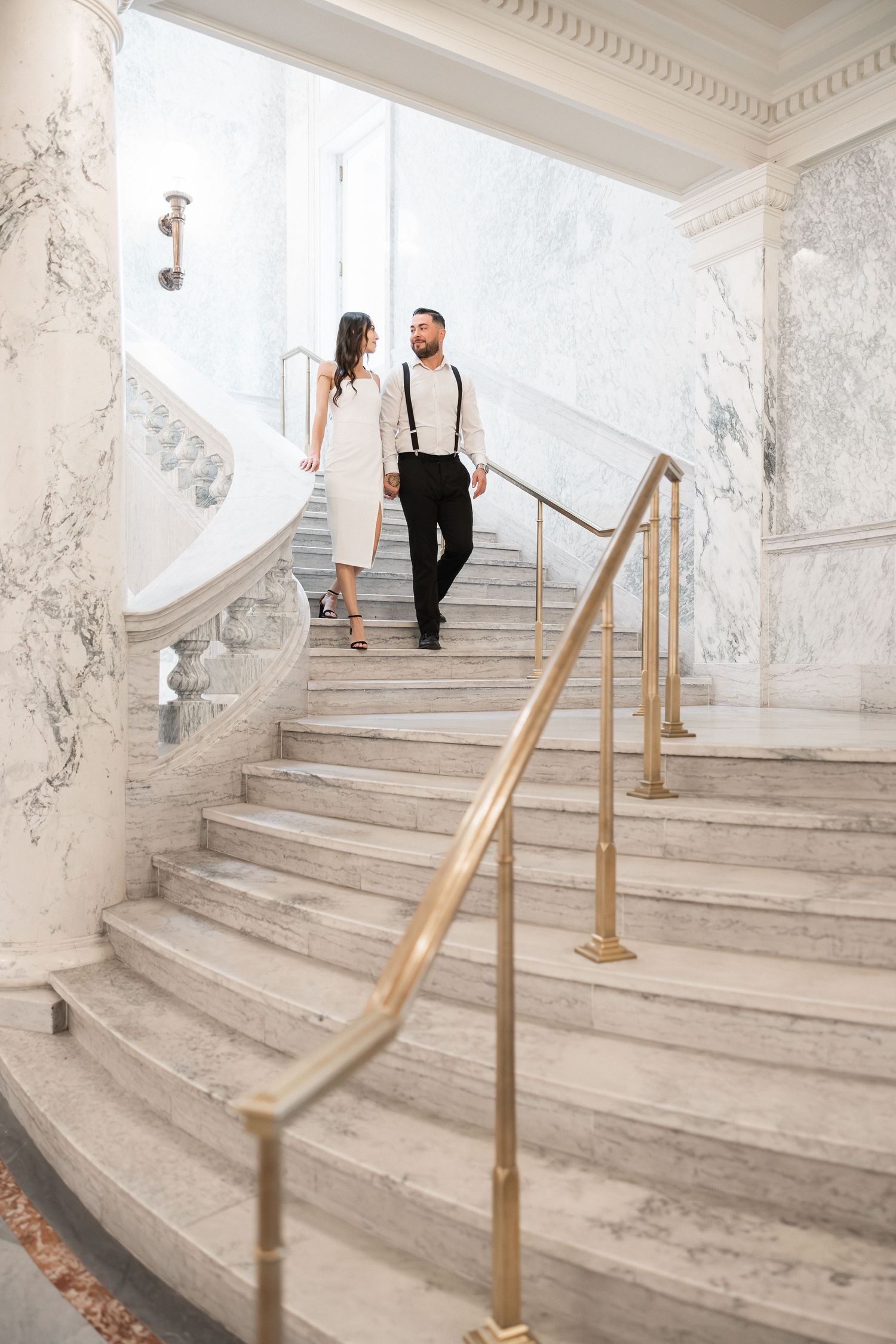 A couple ascends an elegant marble staircase with ornate molding and brass handrails in a luxurious interior.