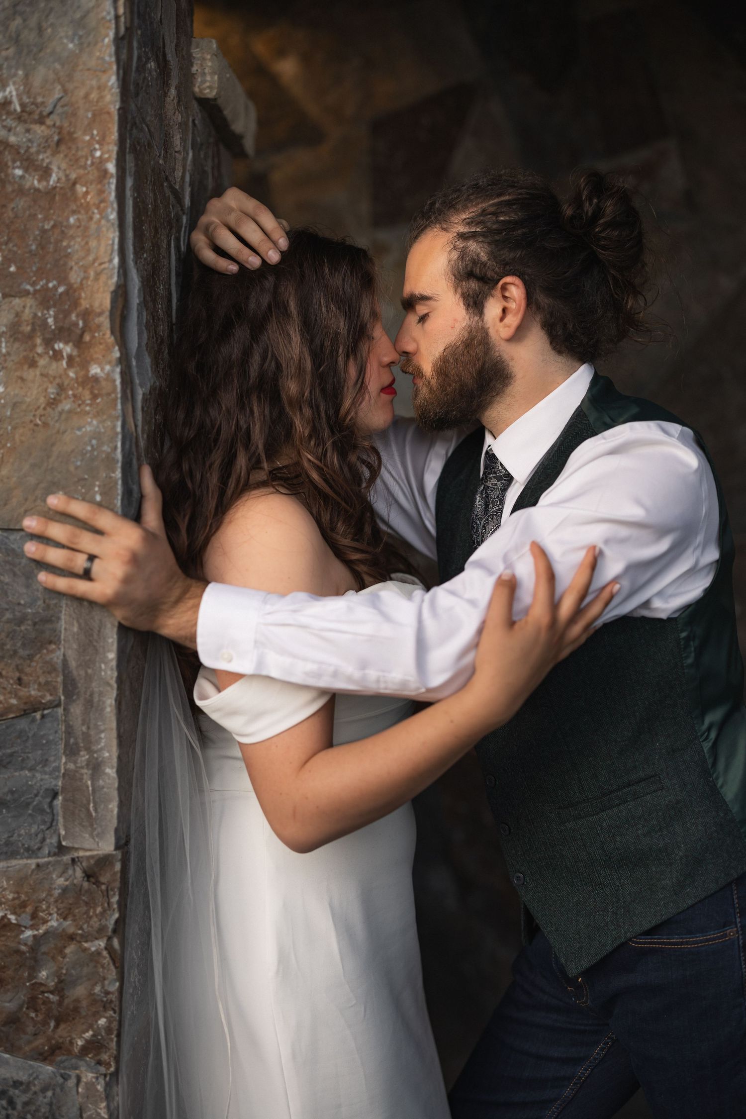 Romantic wedding couple embracing against rustic stone wall in dramatic lighting.