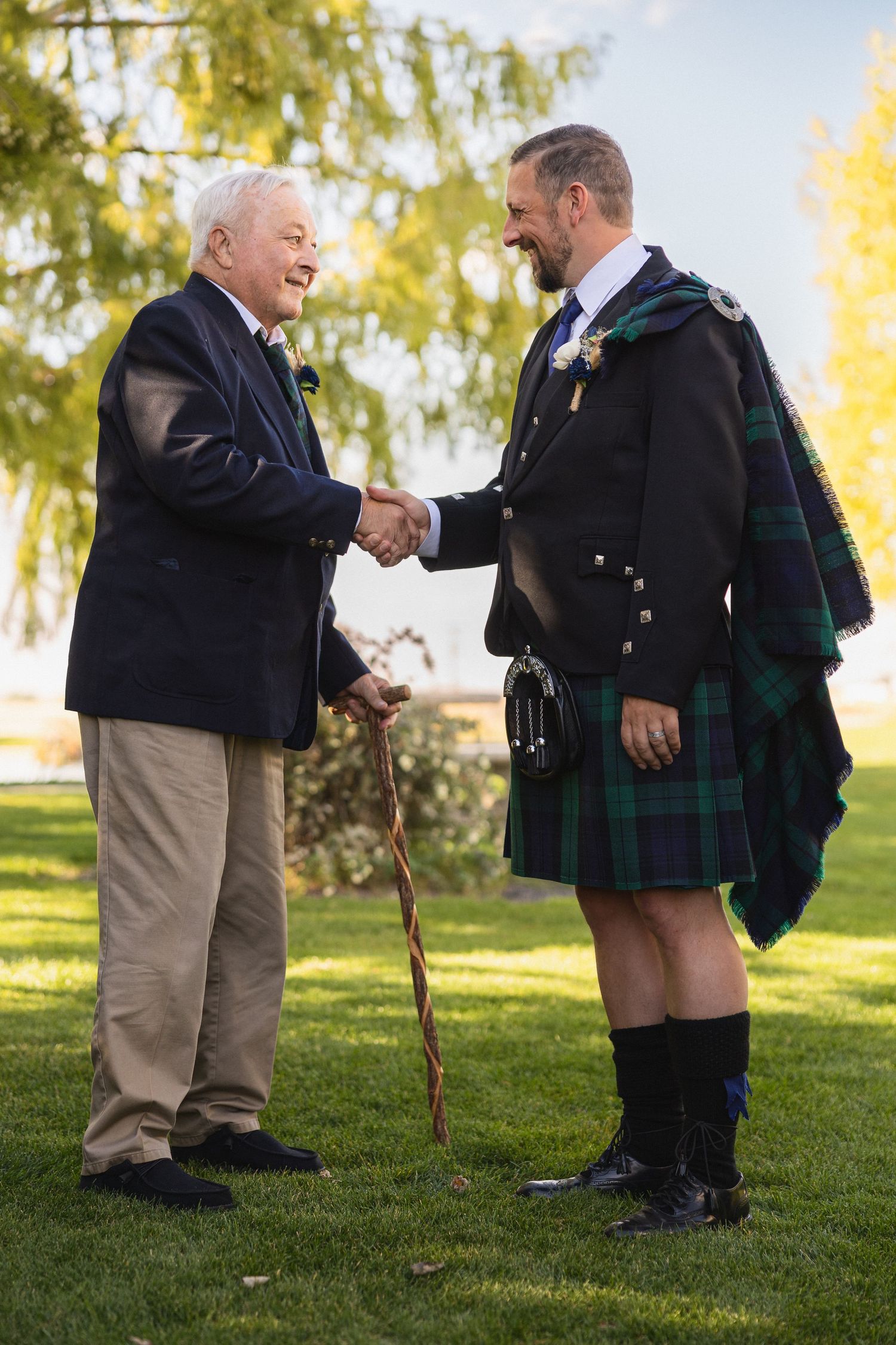 Two people share a handshake outdoors, one wearing a traditional Scottish kilt and the other in casual attire.