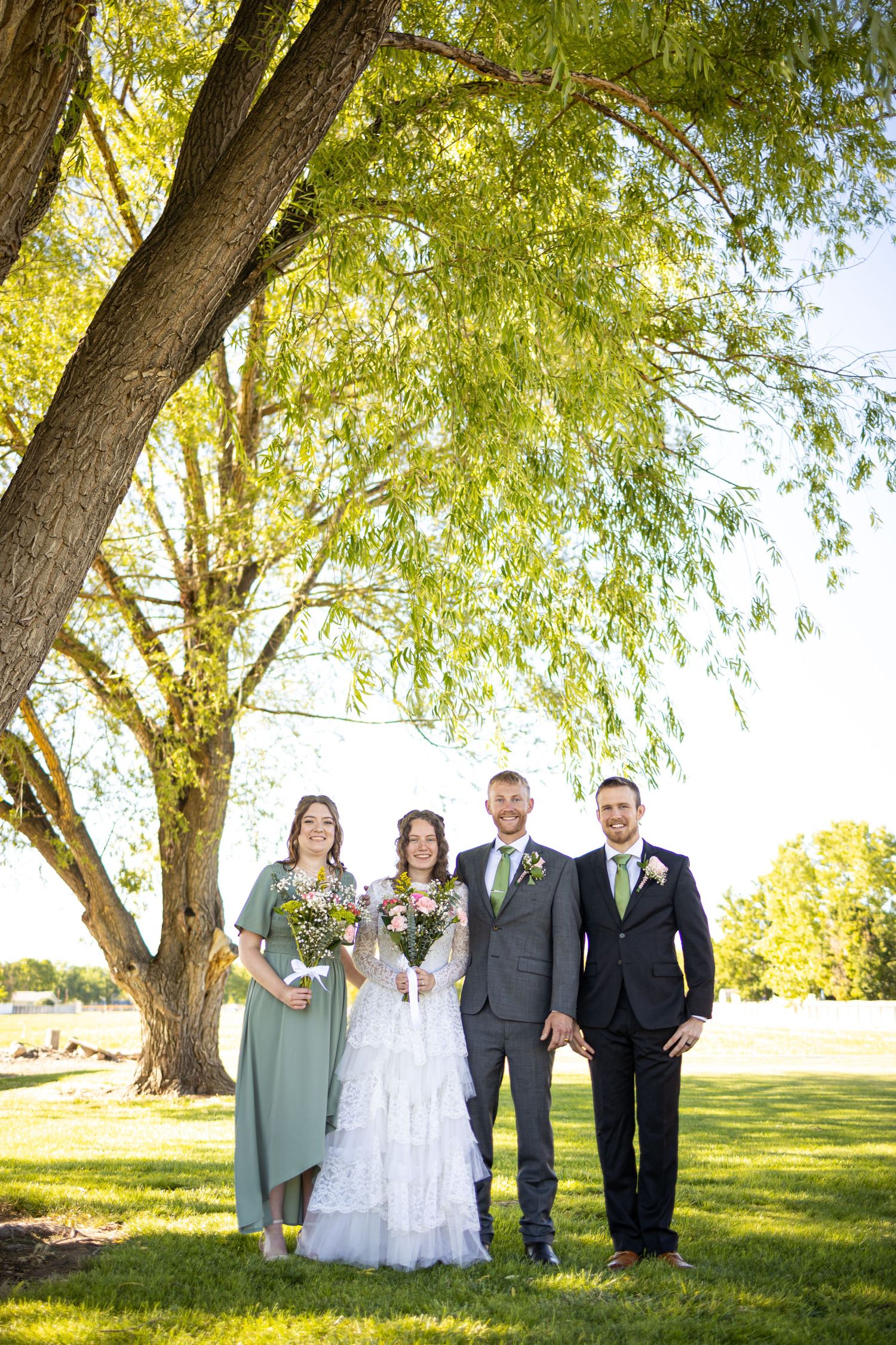 Wedding group poses under large willow tree during outdoor ceremony.