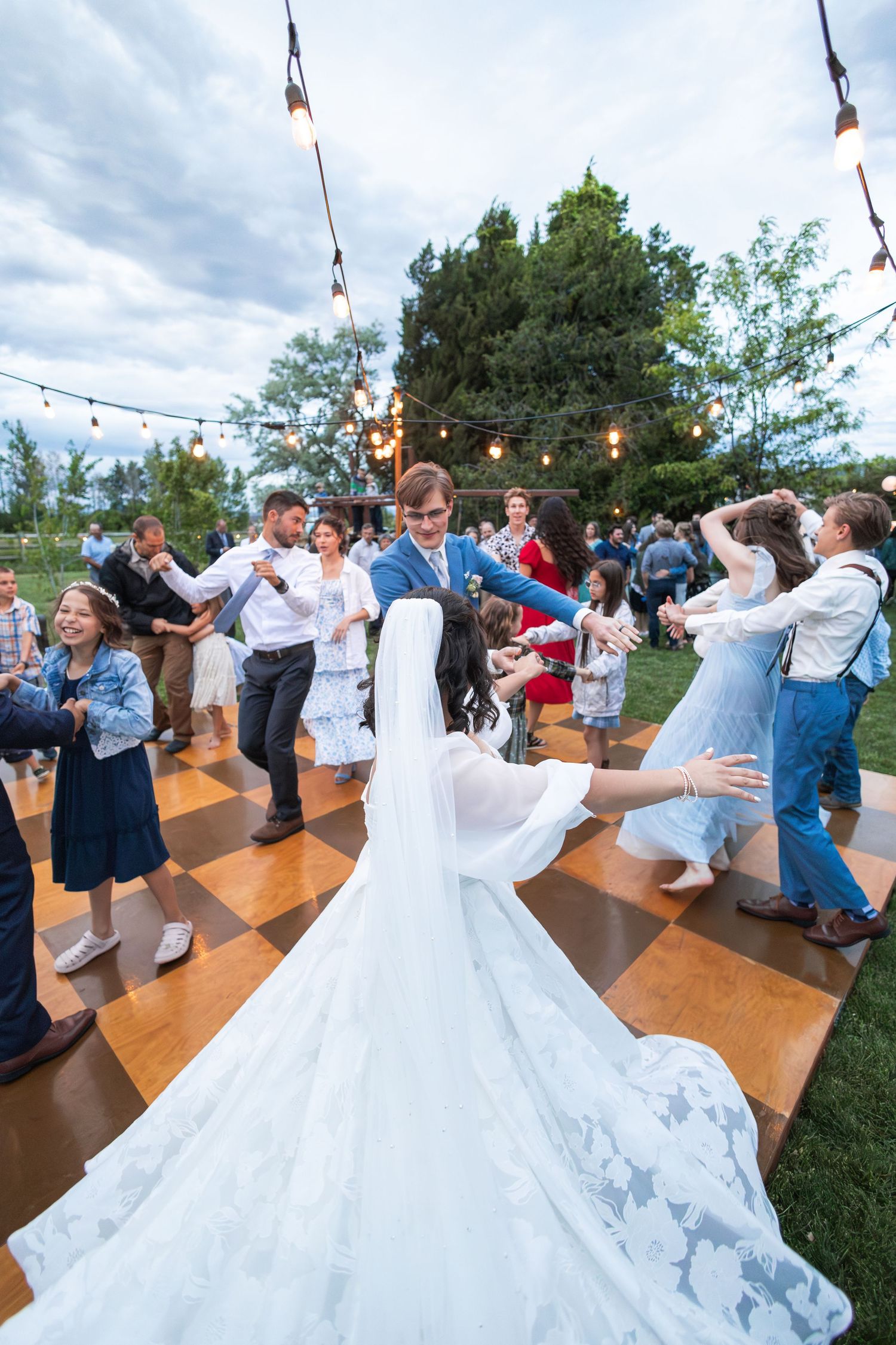 Wedding guests dance together on a wooden dance floor under string lights at an outdoor reception.
