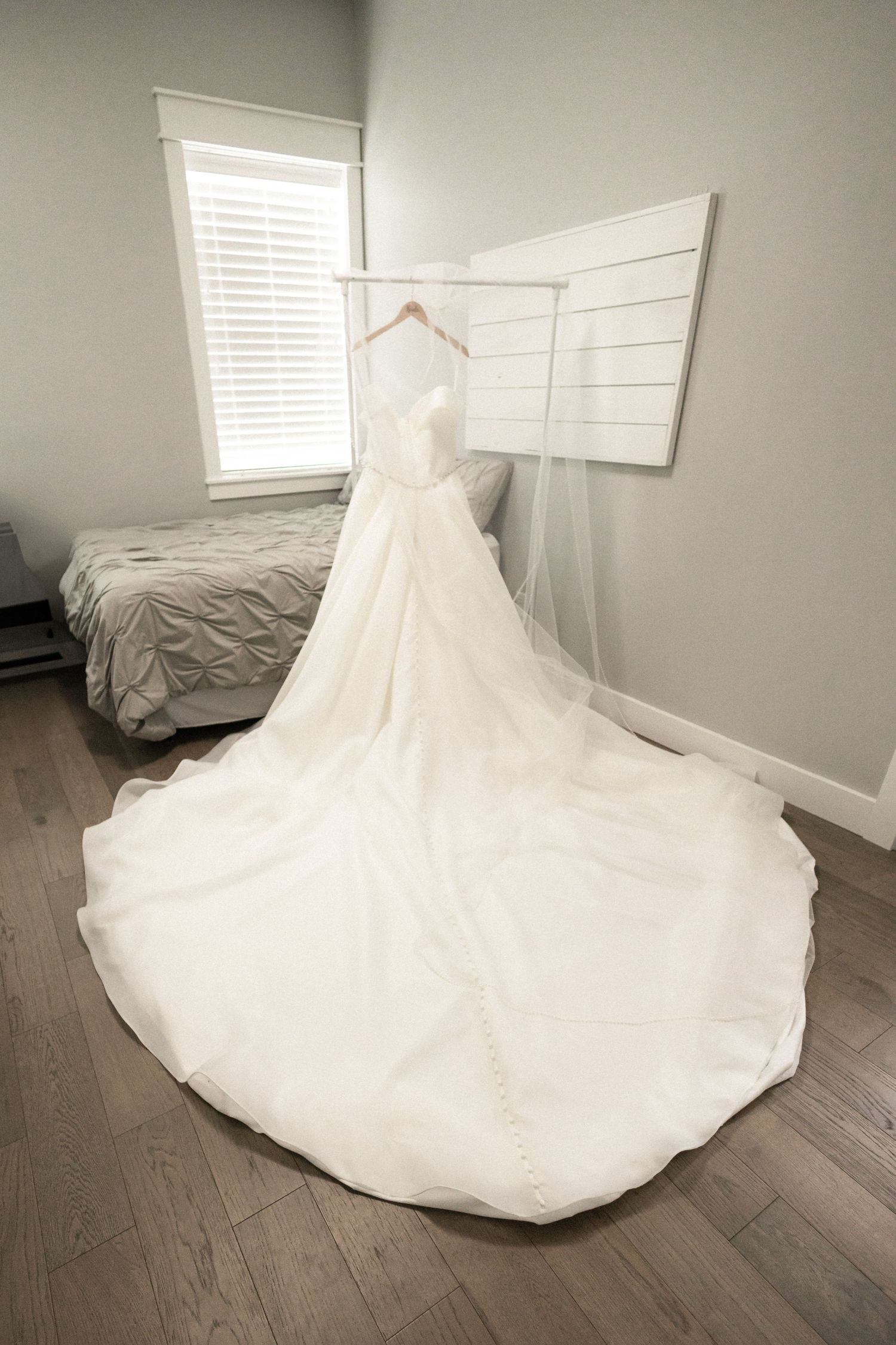 White wedding dress with long train hanging on display in minimal bedroom setting.