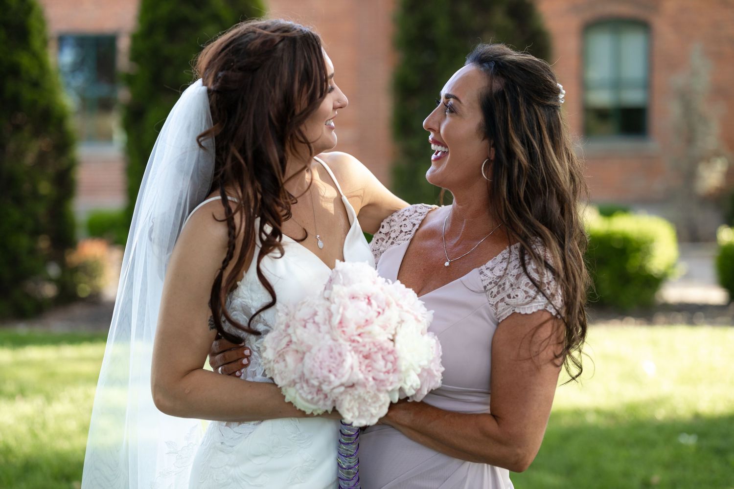 Two people in wedding attire laugh and embrace while holding a pink bouquet of flowers.