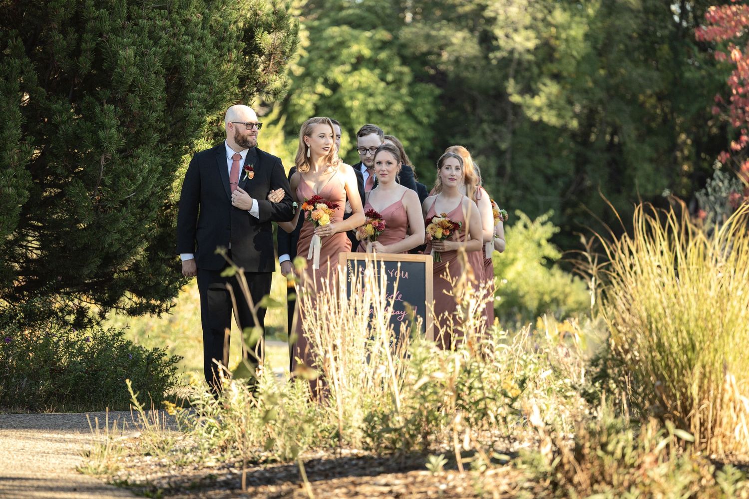 Wedding party walks along an outdoor garden path in formal attire.