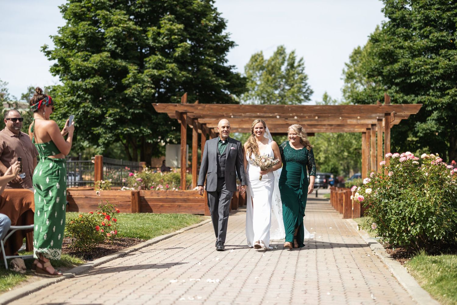 Wedding guests mingle on a brick patio surrounded by lush trees and white gazebos.