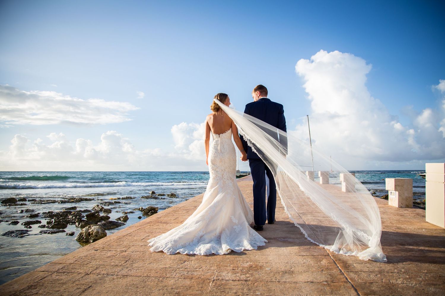 A couple in wedding attire stands on an oceanfront pier as a flowing veil catches the sea breeze against a blue sky.