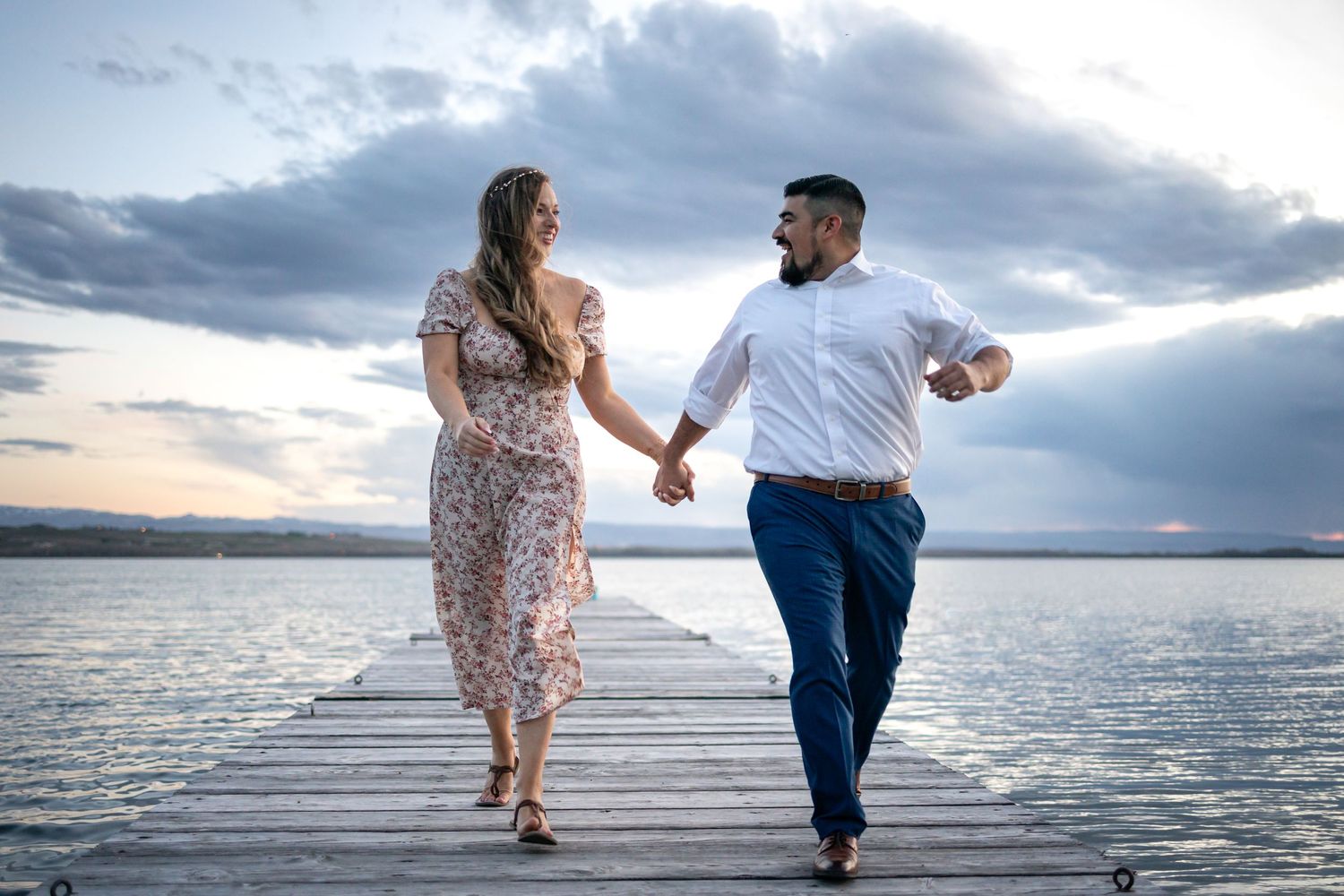 Two people stroll together on a lakeside dock during a cloudy sunset.