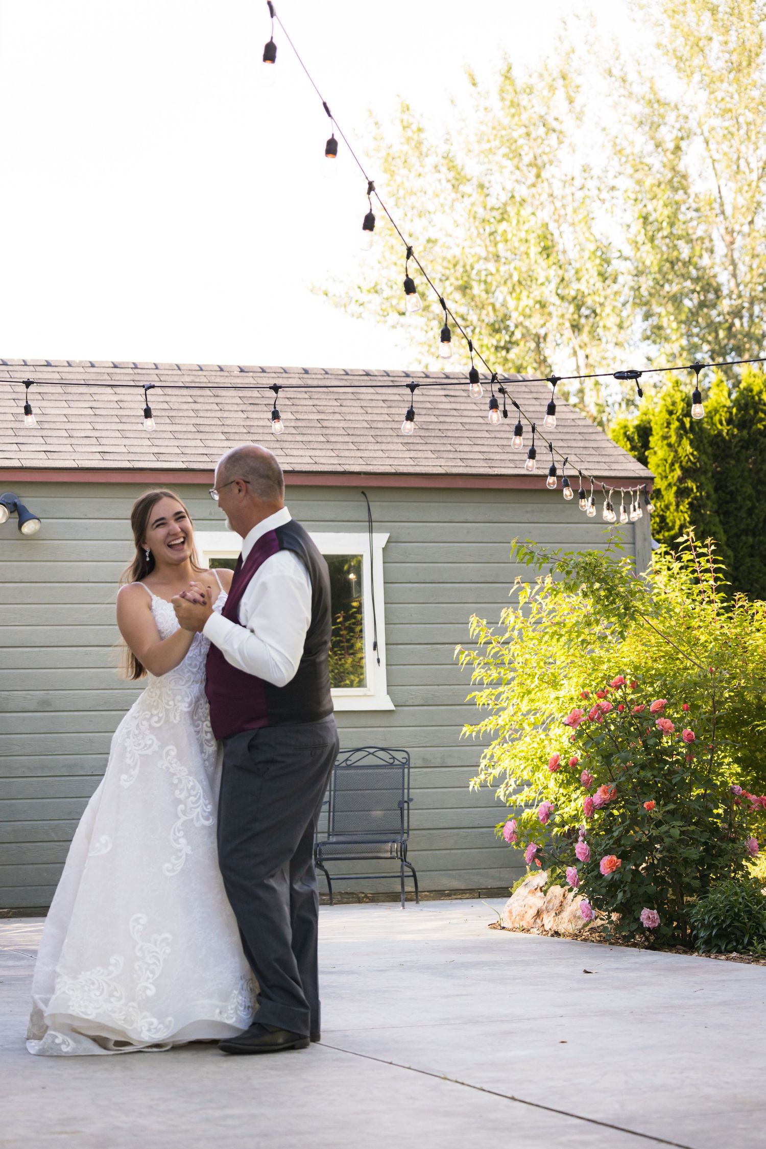 A couple dances under string lights in front of a gray house during a wedding celebration.
