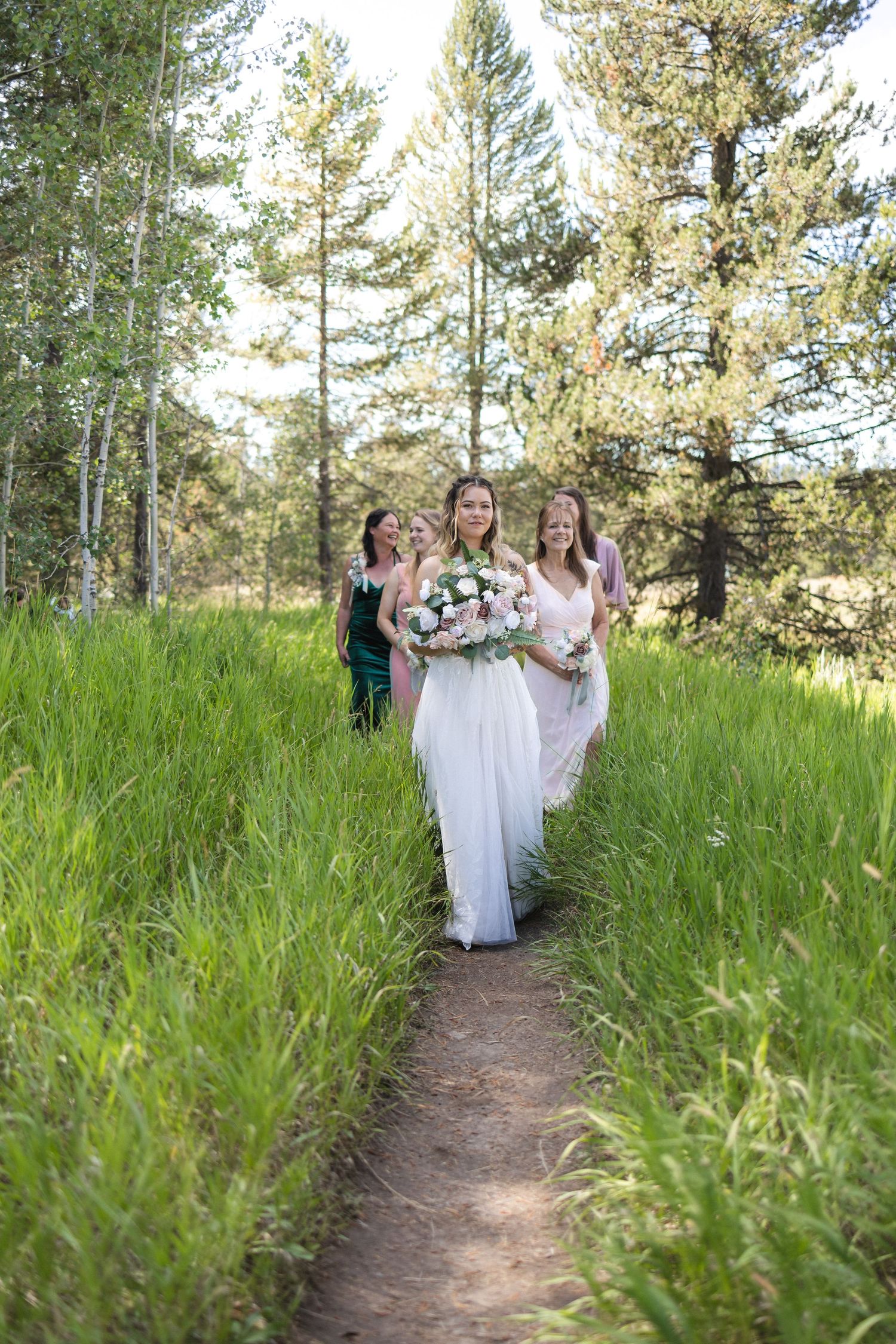 Bridesmaids in white dresses walking along a dirt path through tall grass and pine trees.