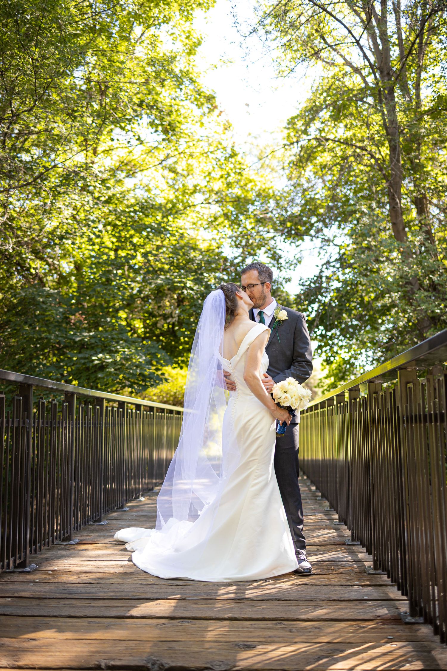 Bride and groom embrace on rustic wooden bridge surrounded by lush summer foliage.