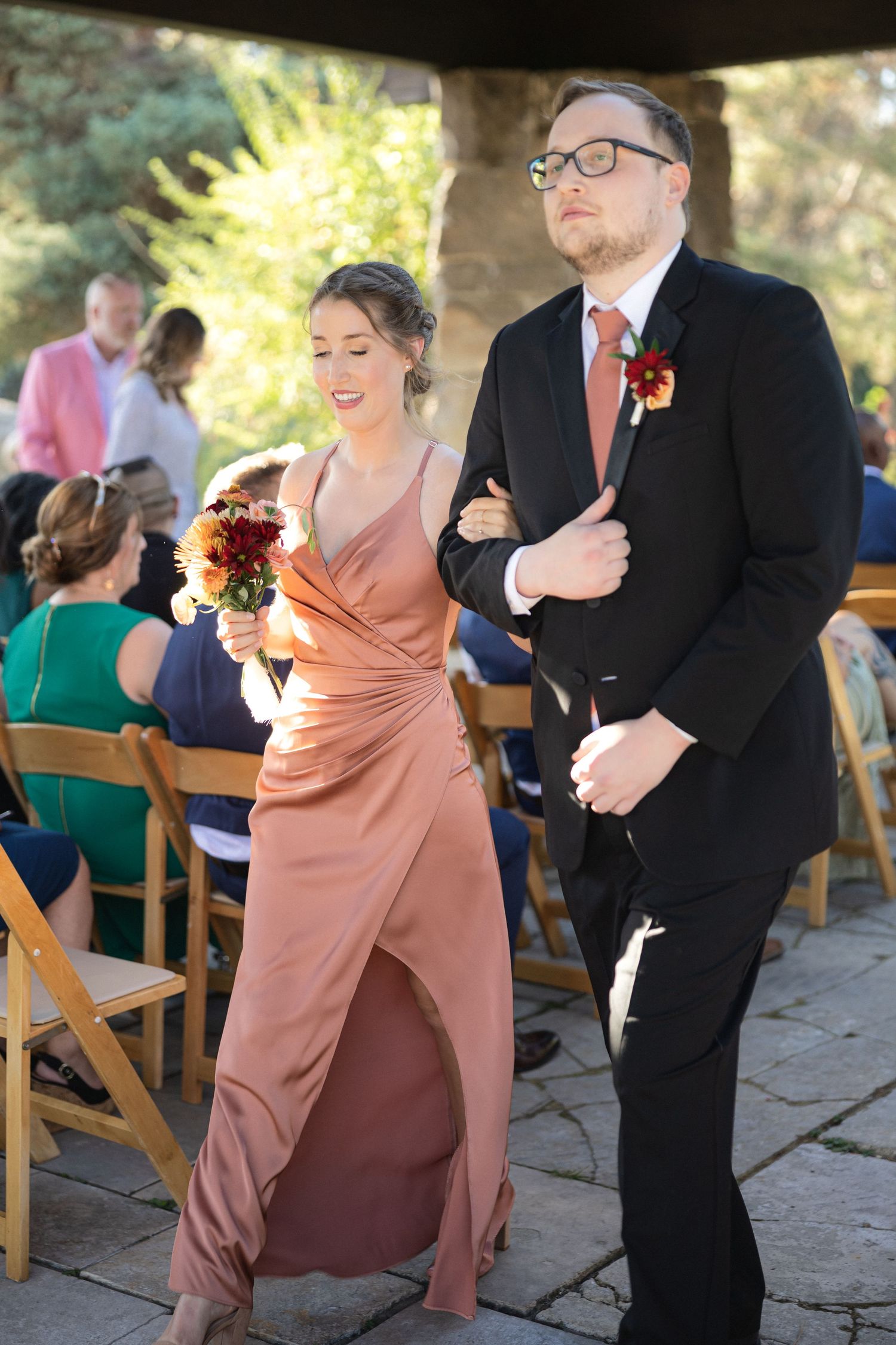 Wedding party member in rose gown walks arm in arm with groomsman during ceremony processional.