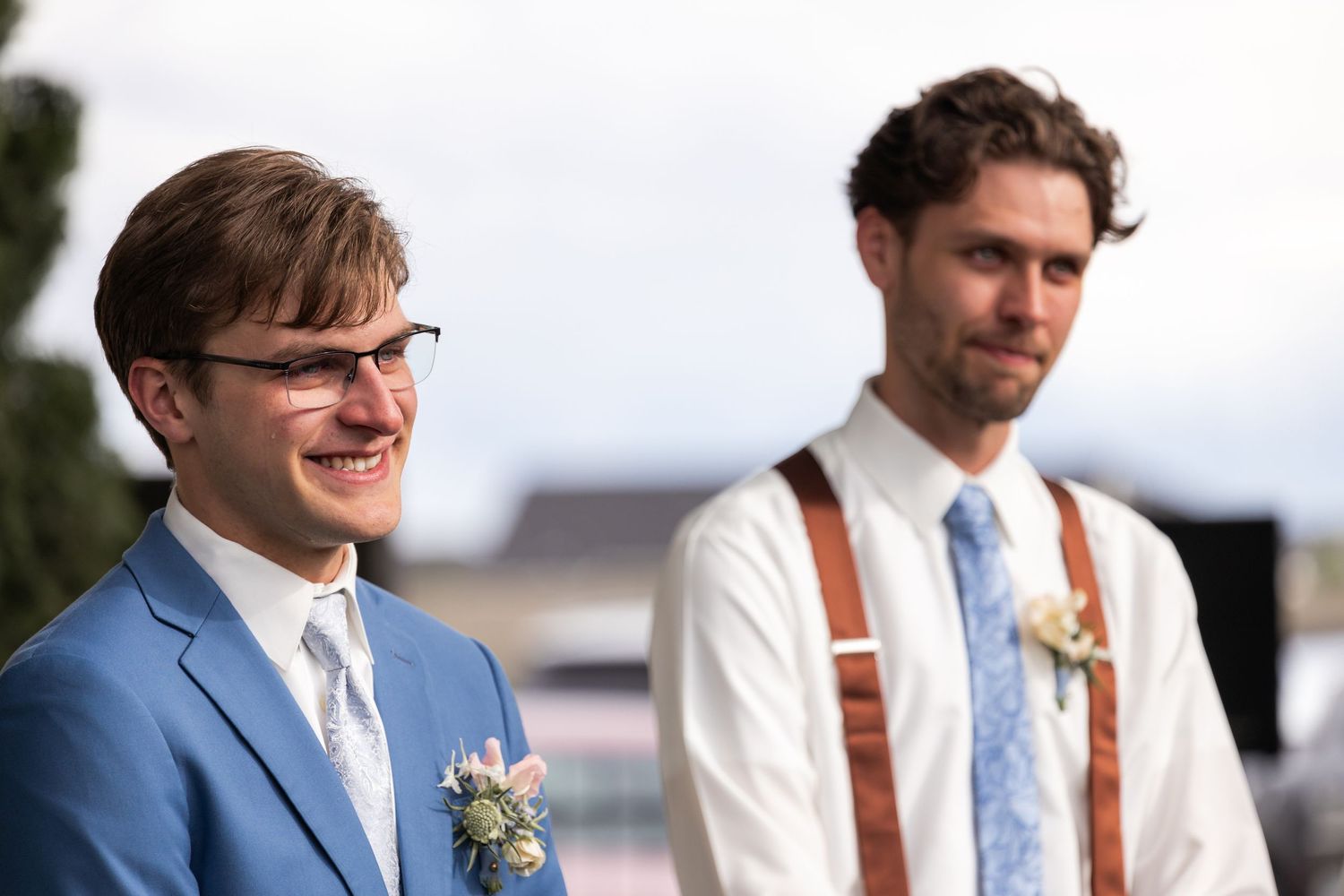 Groom in blue suit and best man in suspenders share emotional moment during outdoor ceremony.