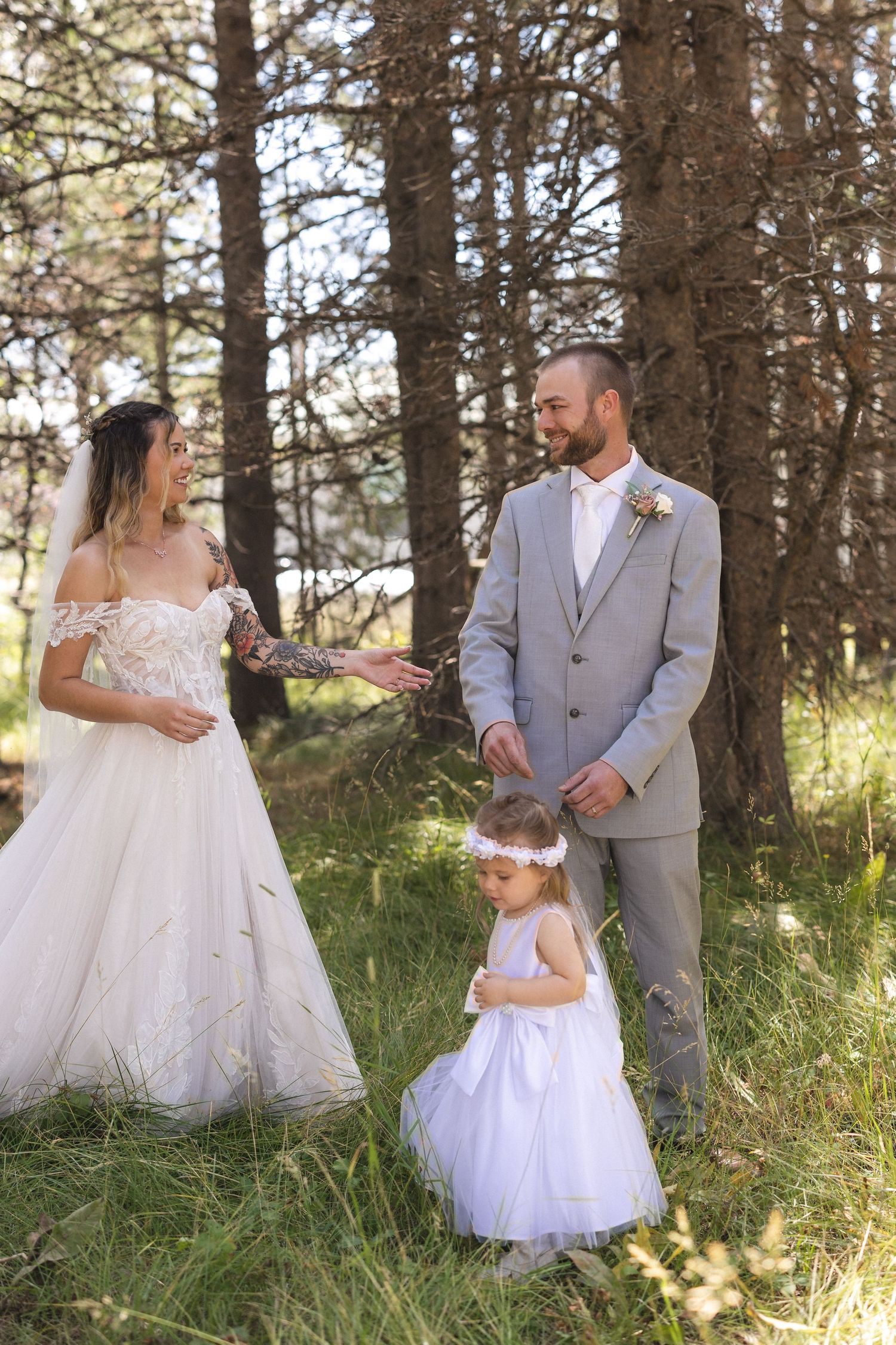 Wedding family portrait in forest with bride, groom in gray suit, and flower girl in white dress.