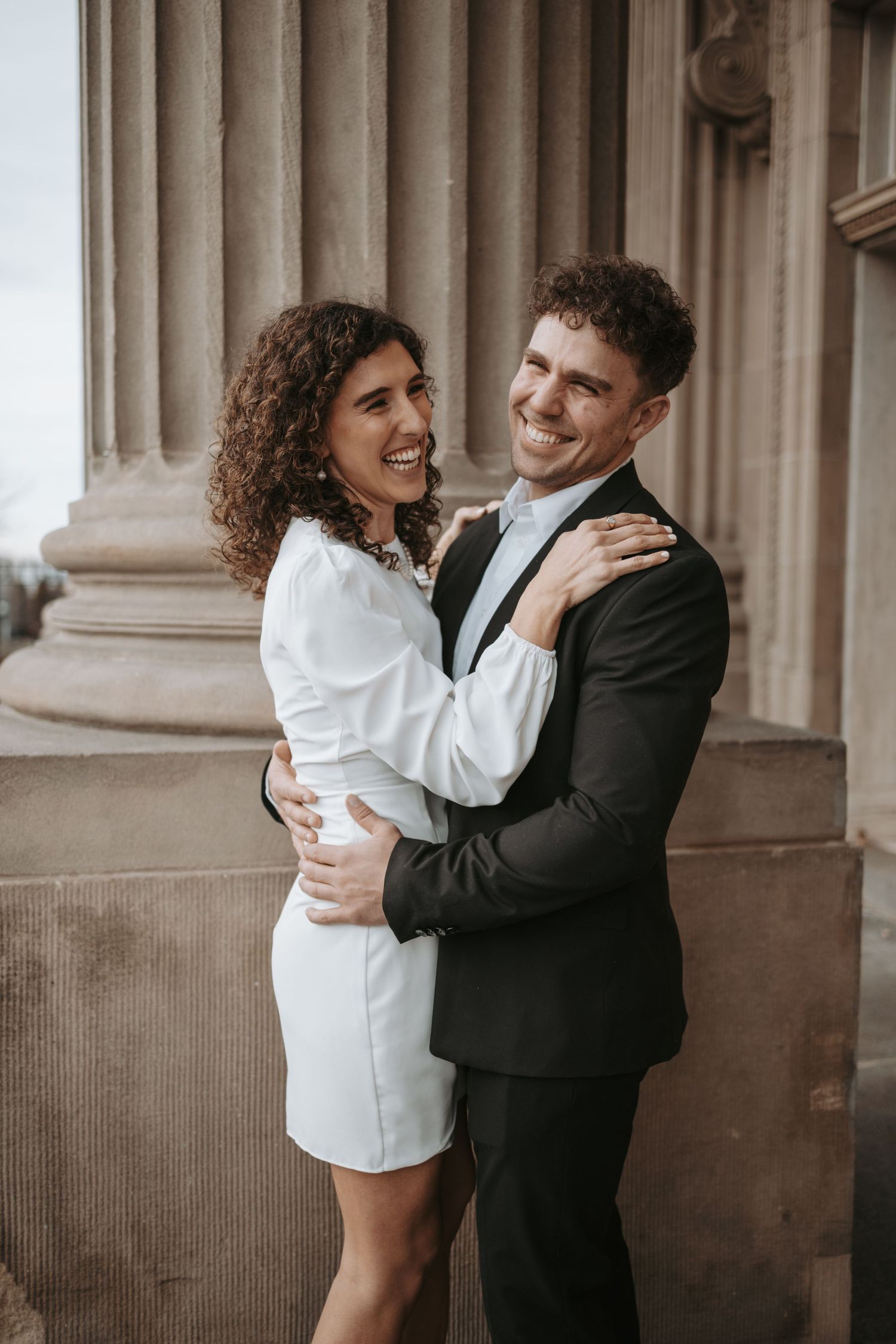A happy couple in formal wear embrace while posing between elegant stone columns.