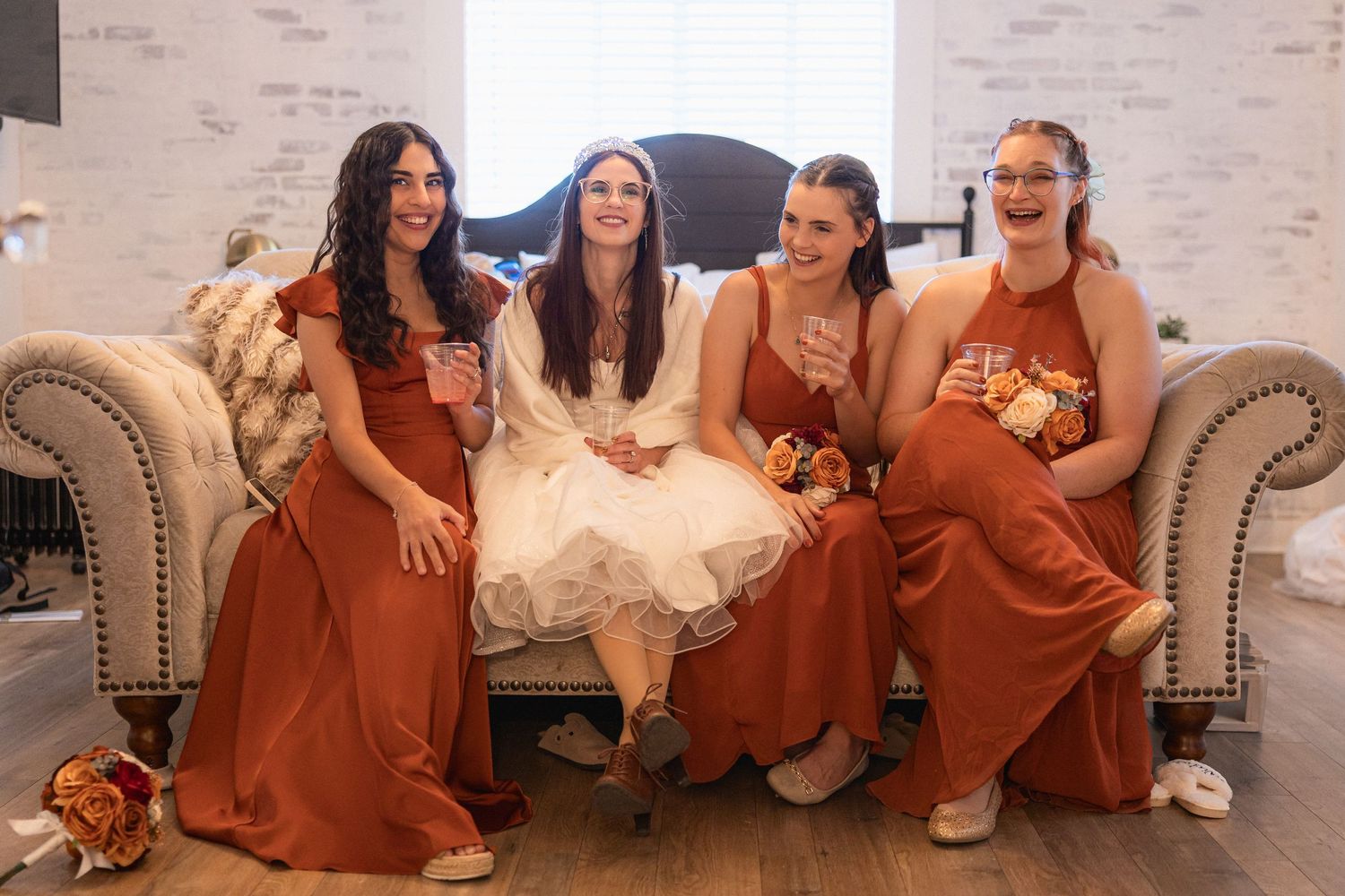 Wedding party seated on a beige couch wearing rust-colored bridesmaid dresses.