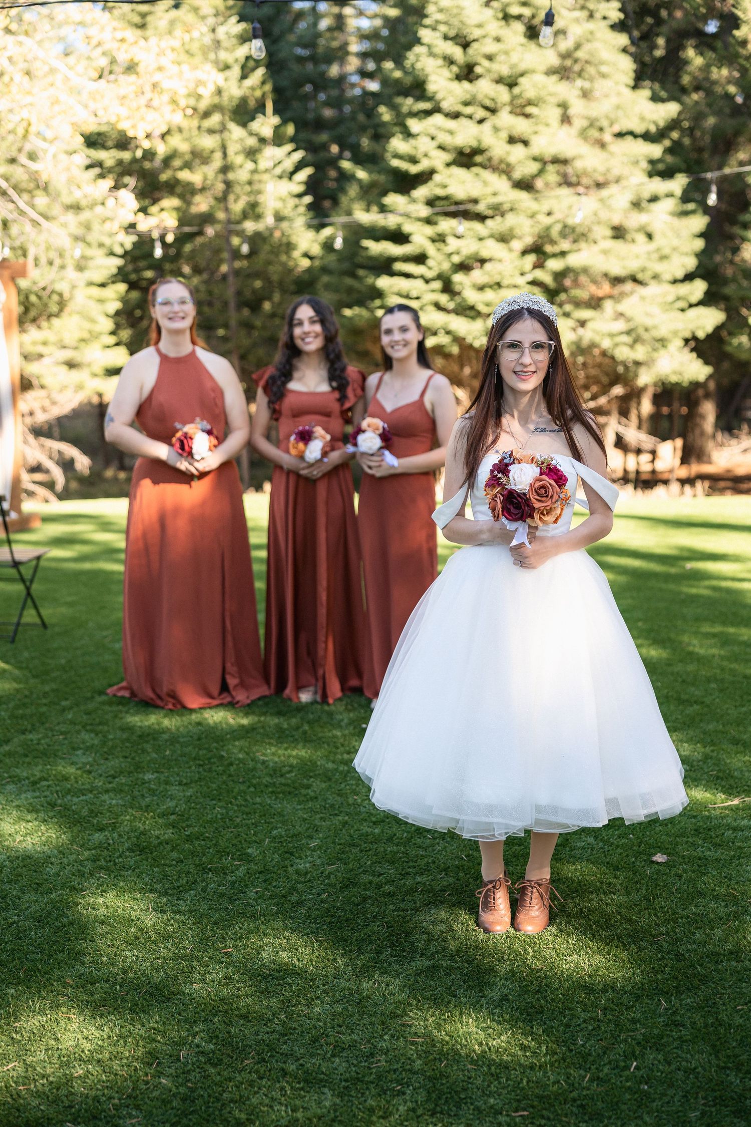A bridal party stands on grass with bridesmaids wearing matching rust-colored dresses.