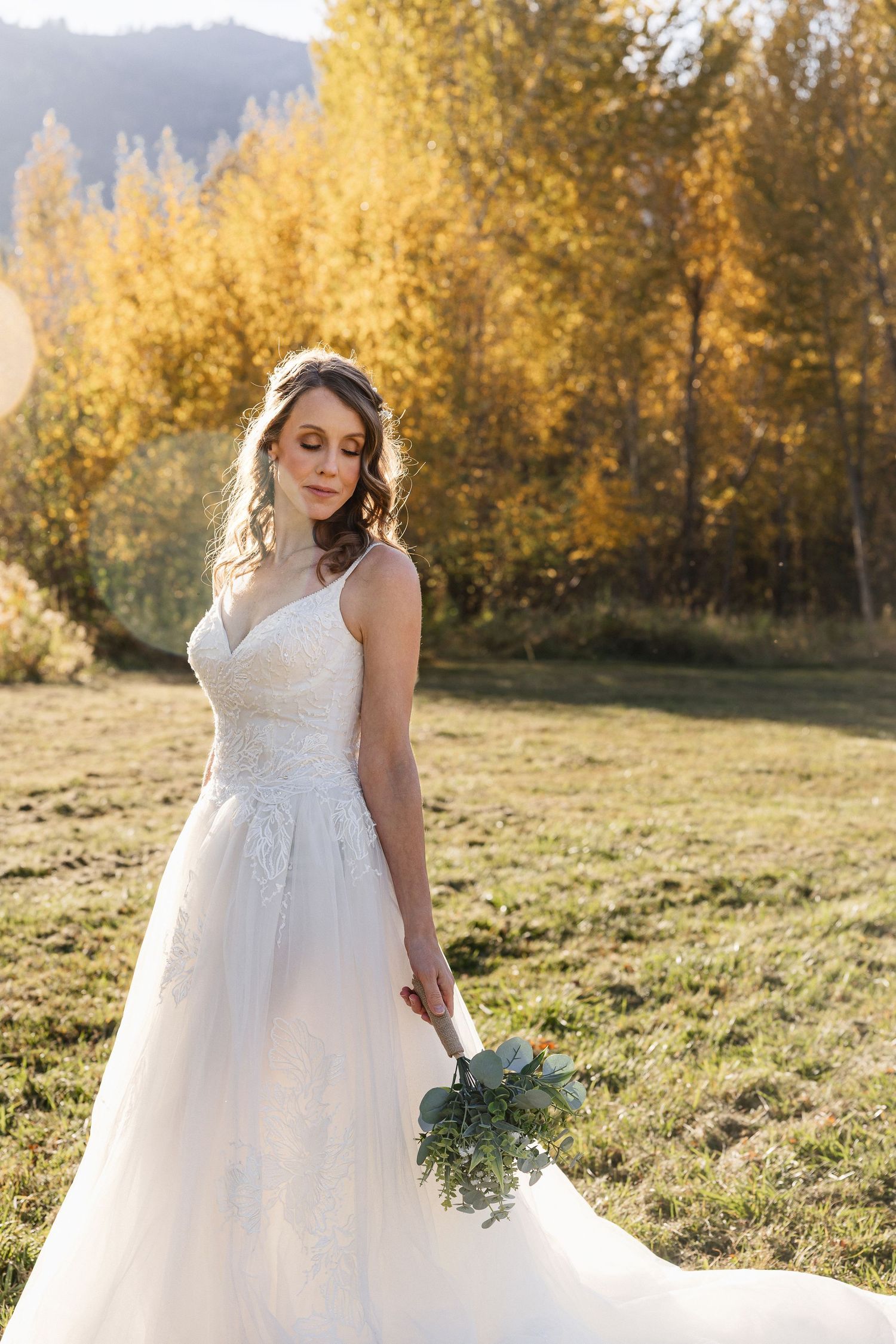A bride in a white lace gown poses with her bouquet against golden autumn foliage.
