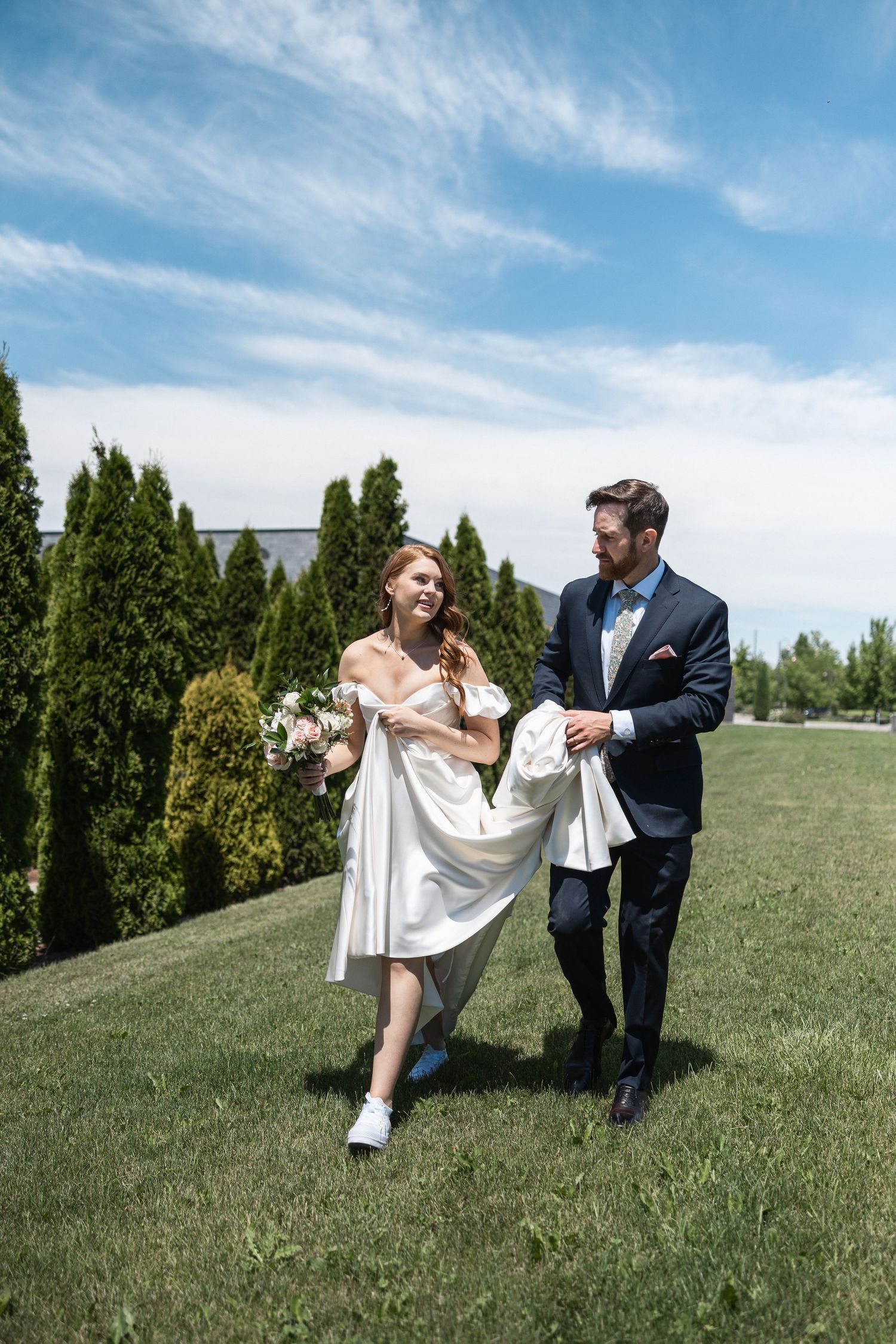 Joyful wedding moment captured on grassy hill with cypress trees and blue sky.
