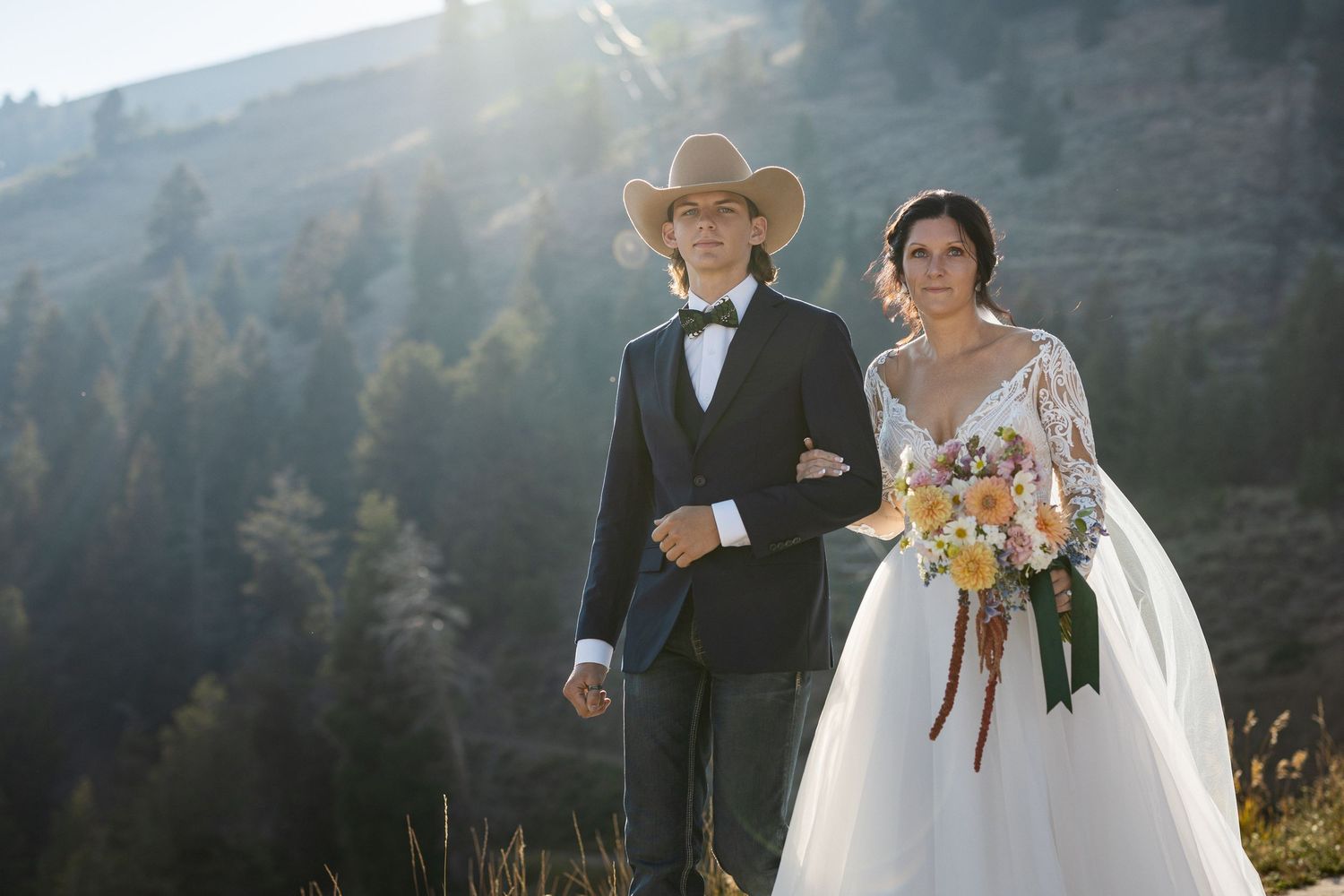 Wedding couple poses on mountain trail with dramatic mountain landscape behind them.