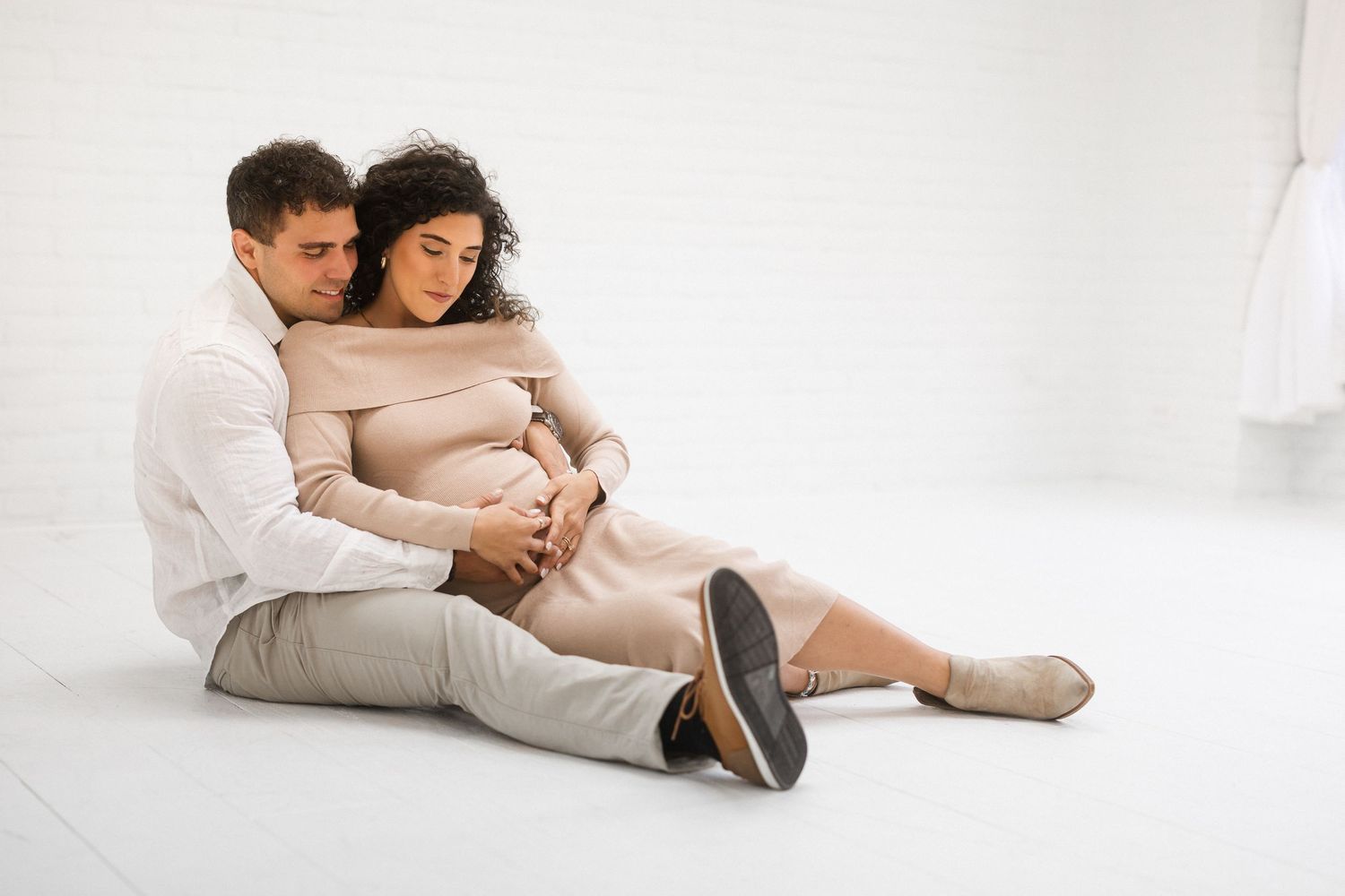 A couple sits together on a white floor during a maternity photo session wearing neutral tones.