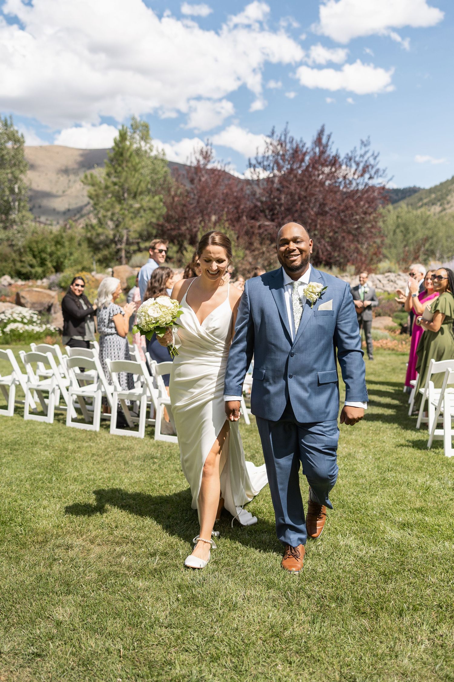Wedding party walking on grass after ceremony with white chairs and mountains in background.