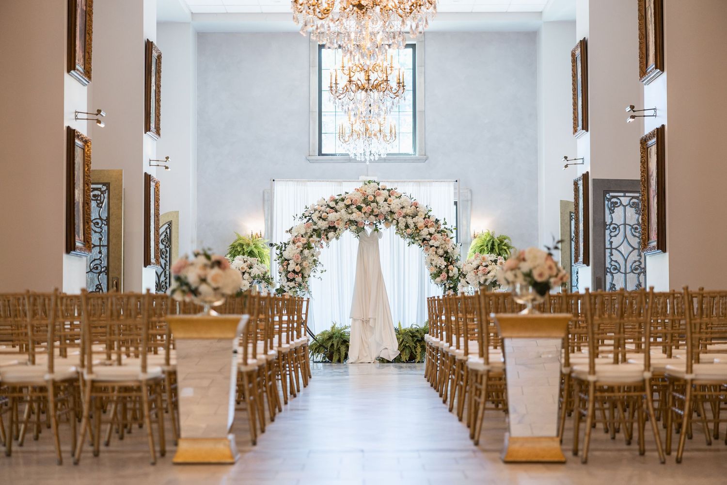 Spacious white wedding venue with crystal chandeliers and wooden chairs arranged in rows for ceremony seating.