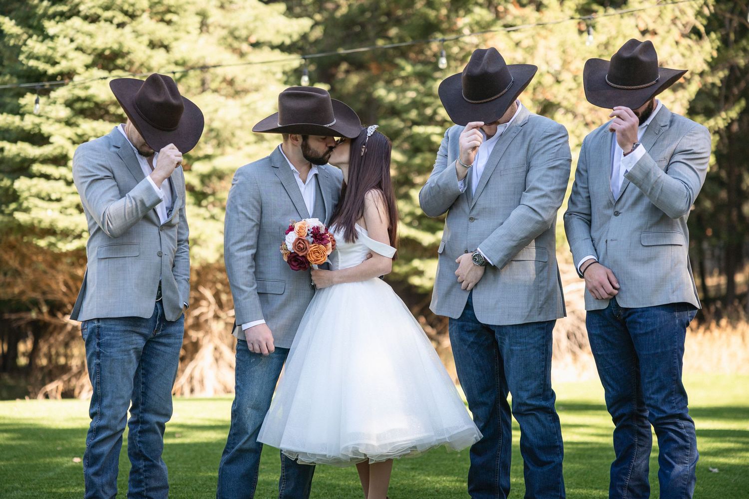 Wedding party shares a lighthearted moment in western-themed attire on sunny lawn.