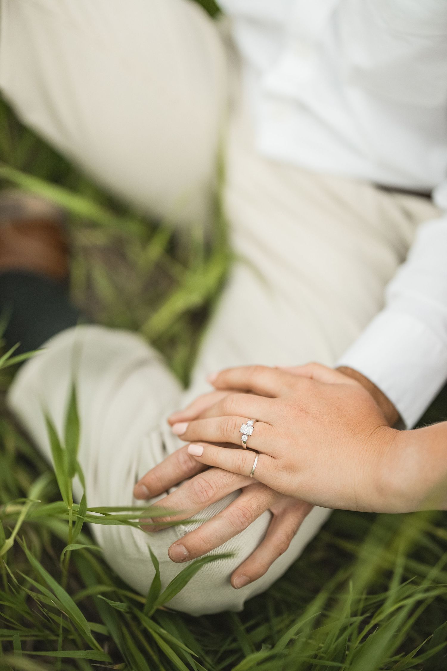 Couple sitting in green grass holding hands, woman wearing a diamond engagement ring, romantic outdoor setting.
