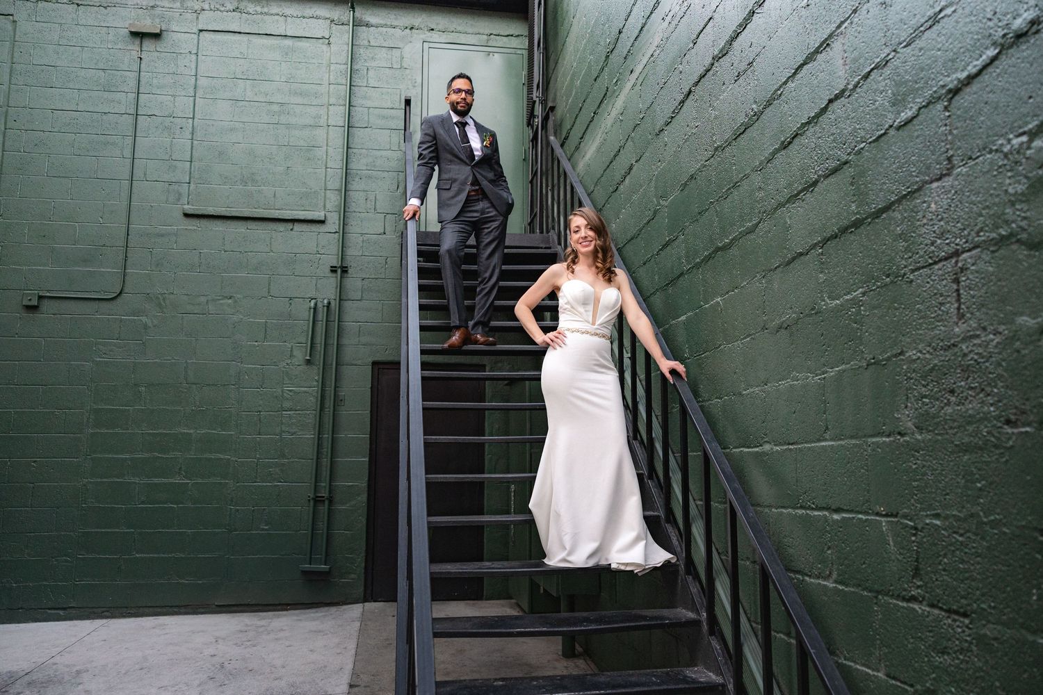Newlyweds stand together on black metal stairs against a green industrial wall.
