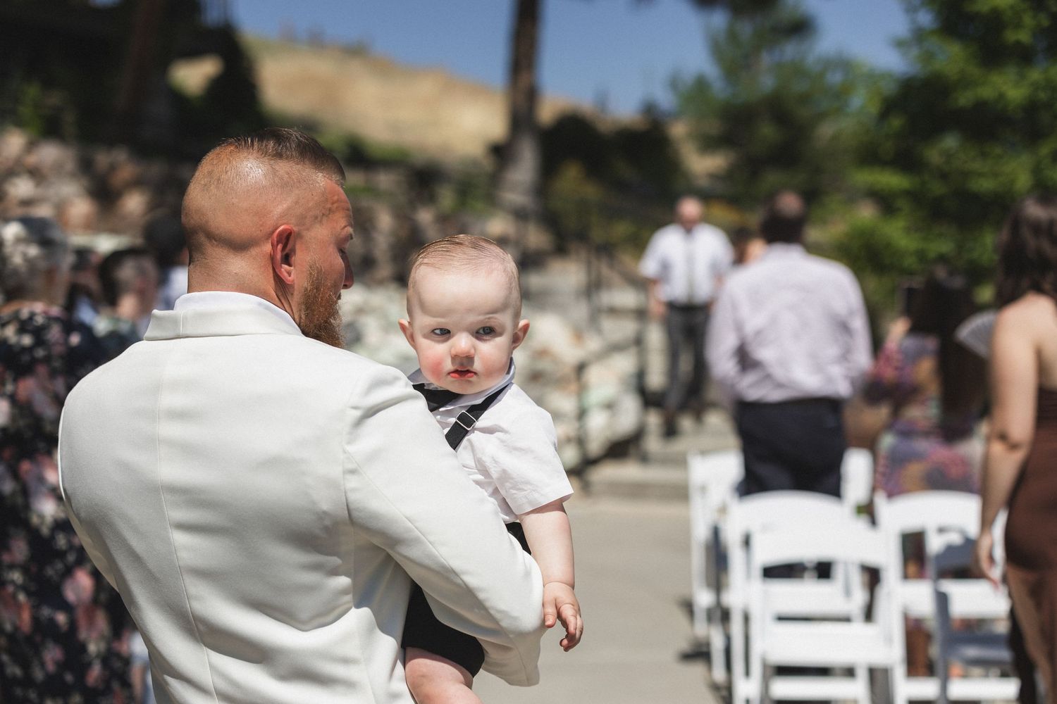 A person in white holds a baby while standing among wedding ceremony guests.