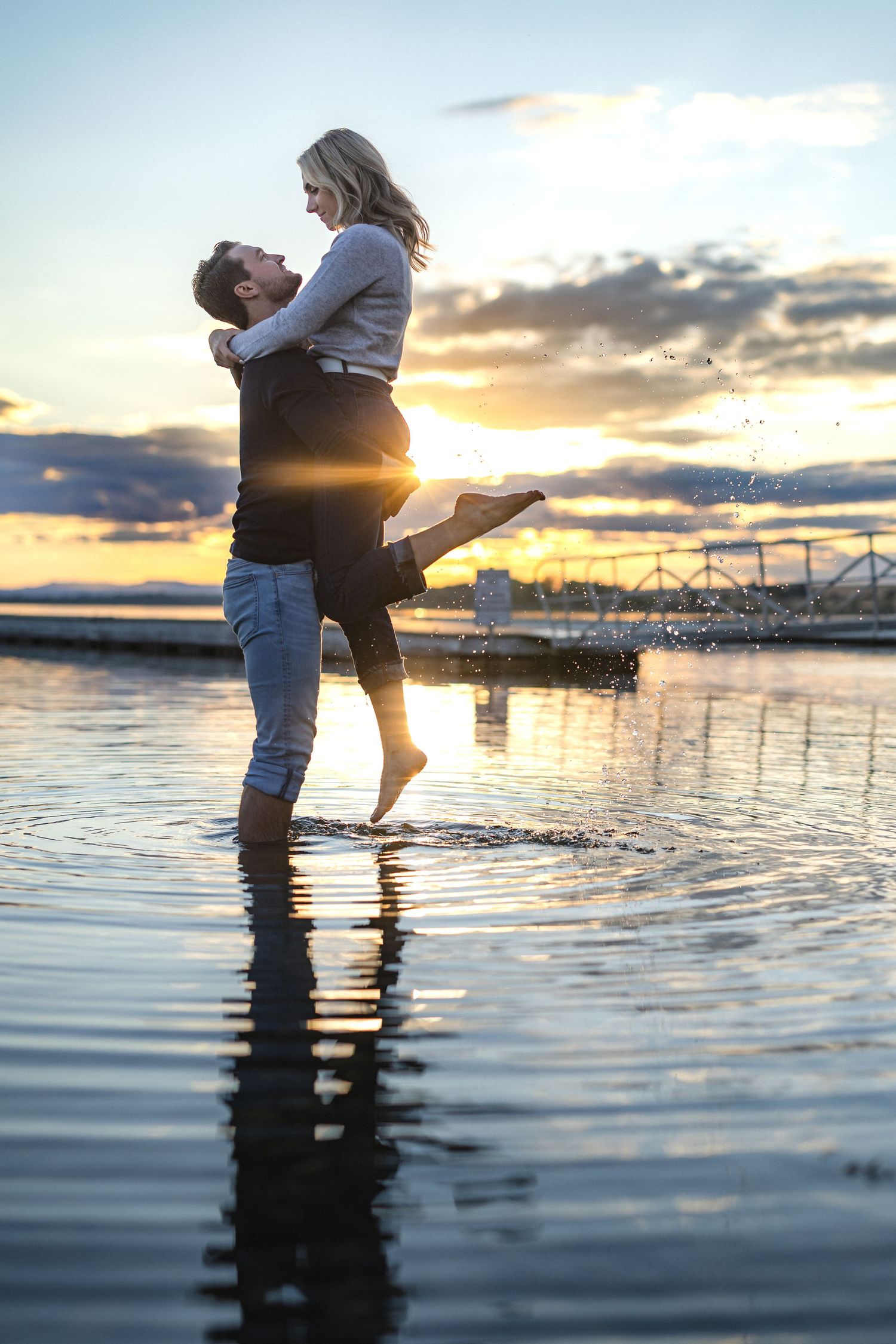 Romantic couple standing in water at sunset with silhouetted dock in distance.