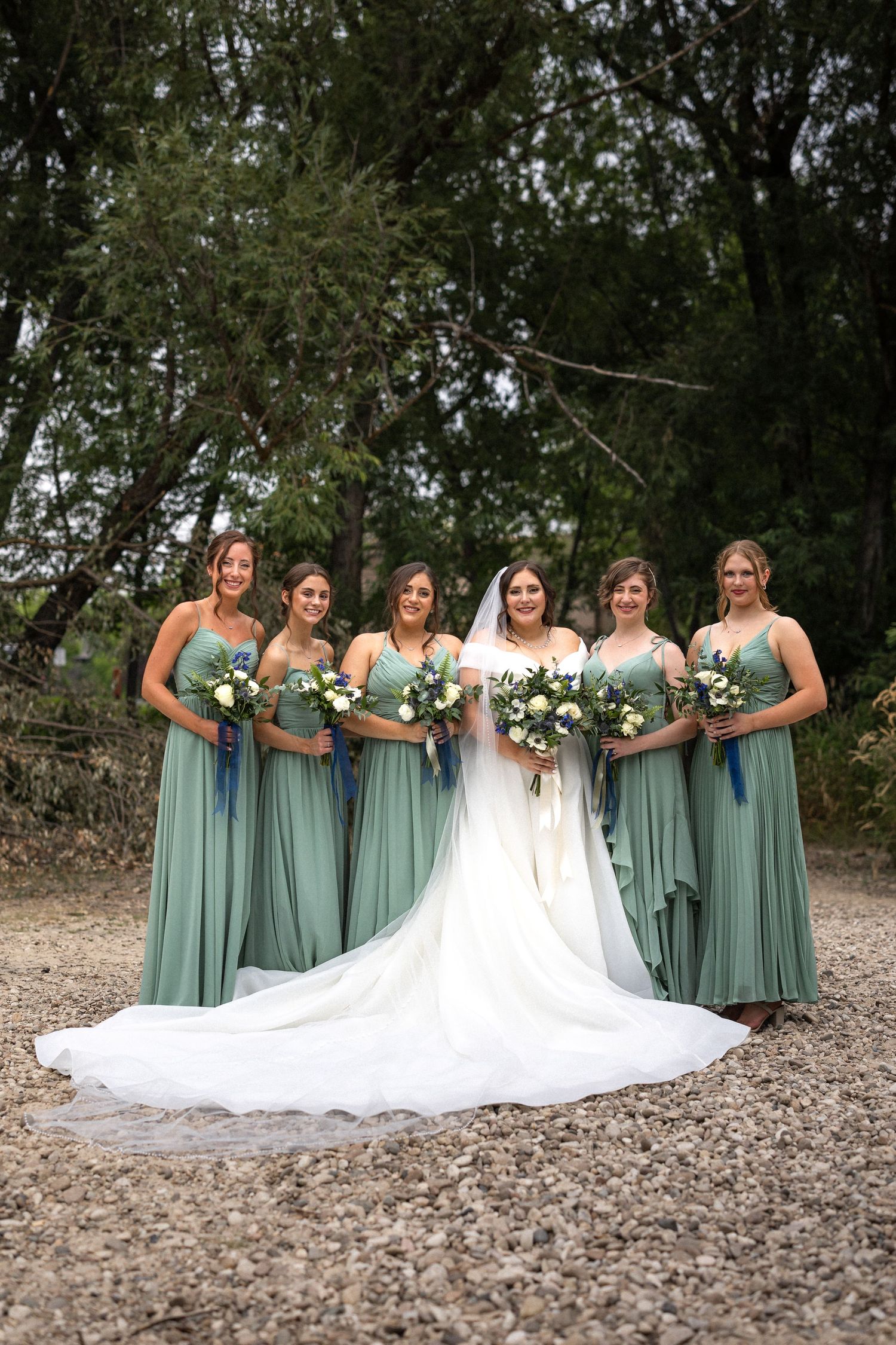 Wedding party portrait showing bride in white dress with train alongside bridesmaids in sage green gowns.