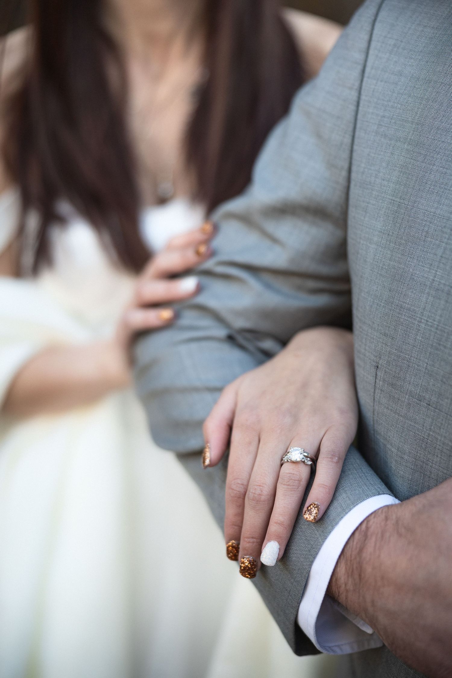 Close-up of hands showing engagement ring with light gray suit and white dress details.