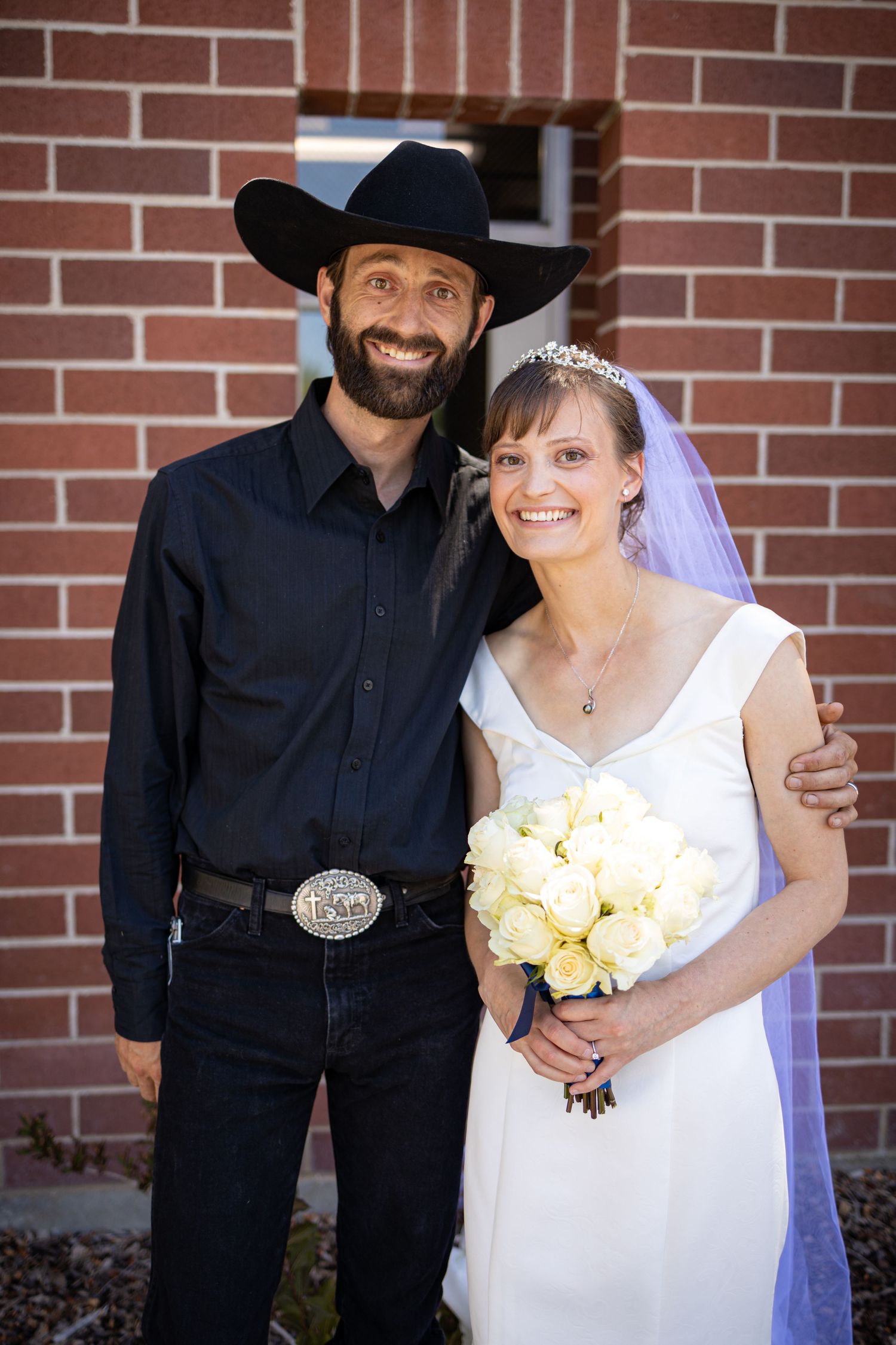 Wedding couple in western attire poses together against brick wall.