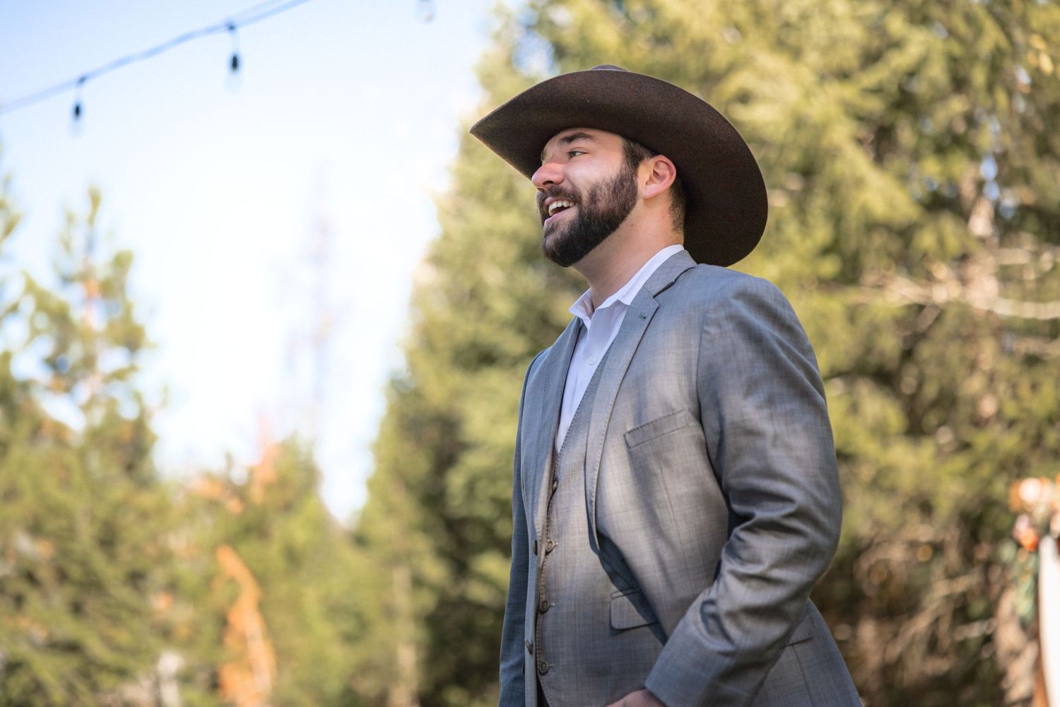 Groom wearing cowboy hat and grey suit smiling in outdoor setting.