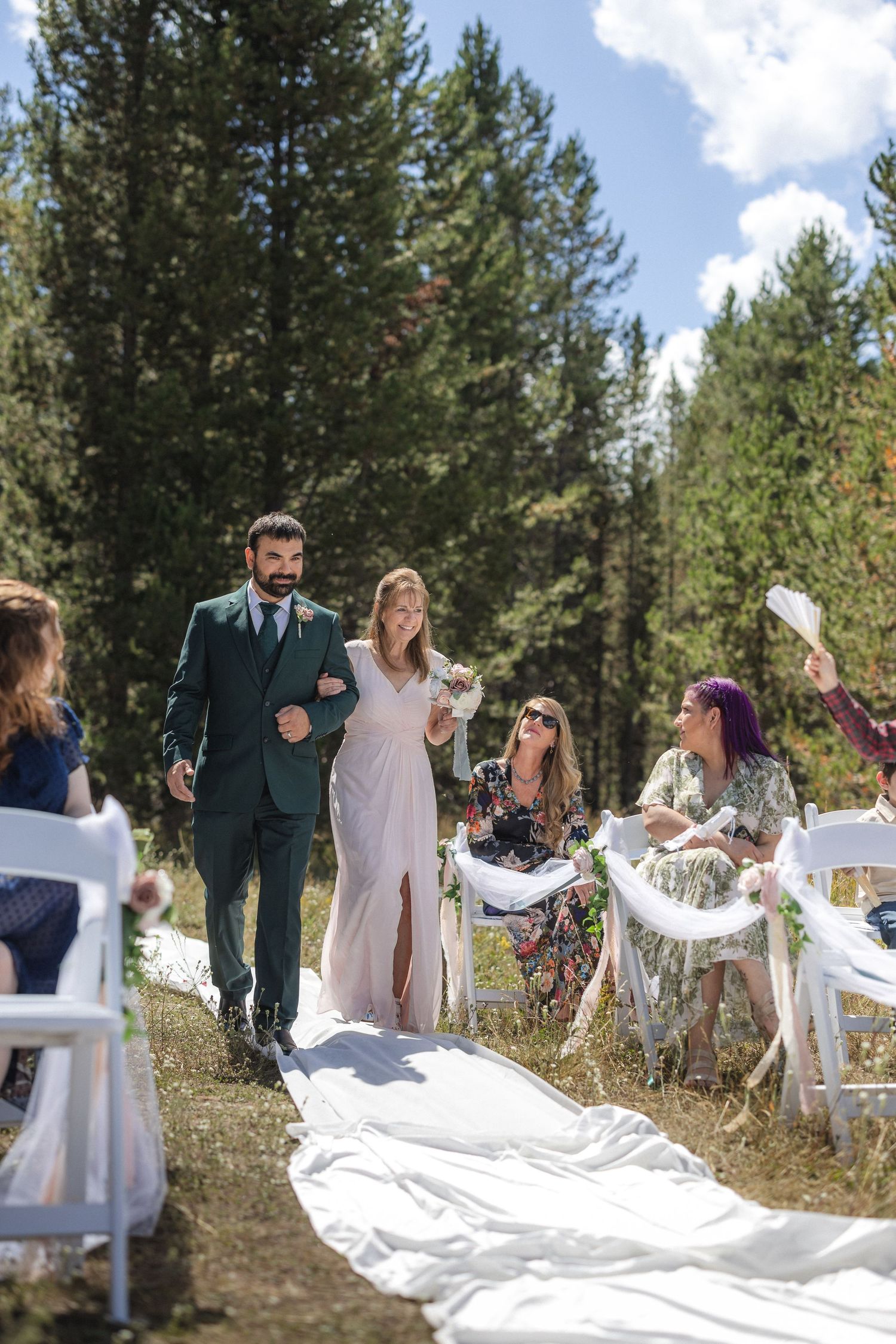 Couple walking down outdoor wedding aisle while guests watch from white chairs.