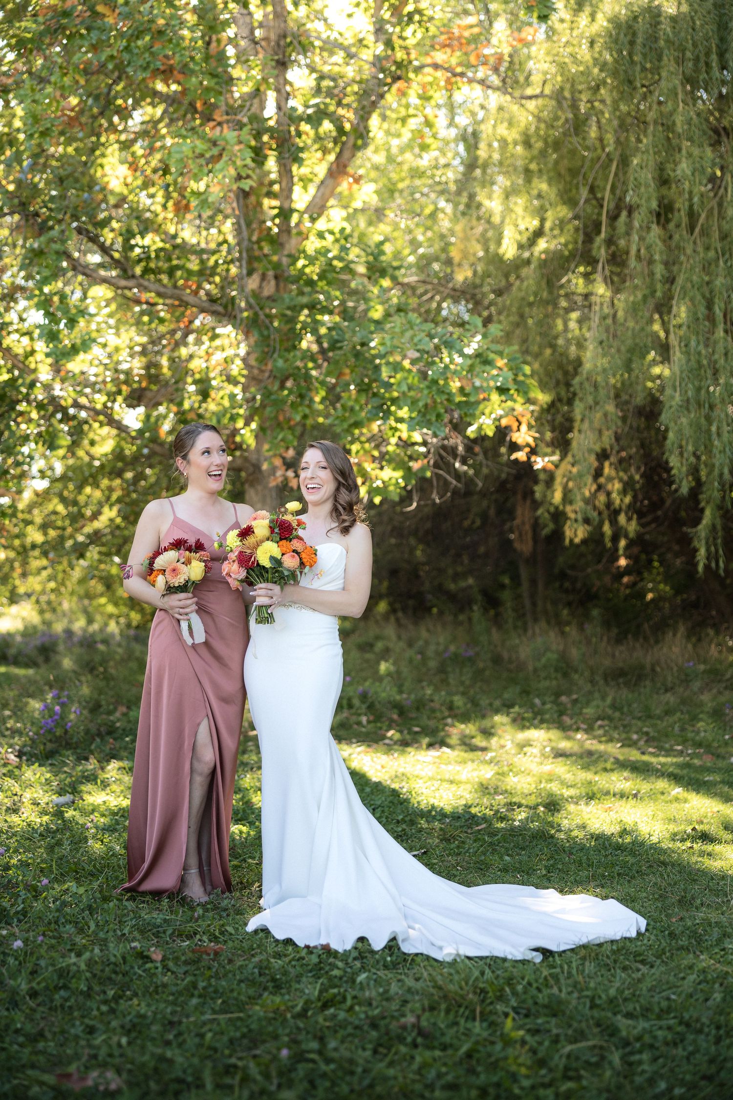 Wedding party poses outdoors in dusty rose dresses against a willow tree backdrop.