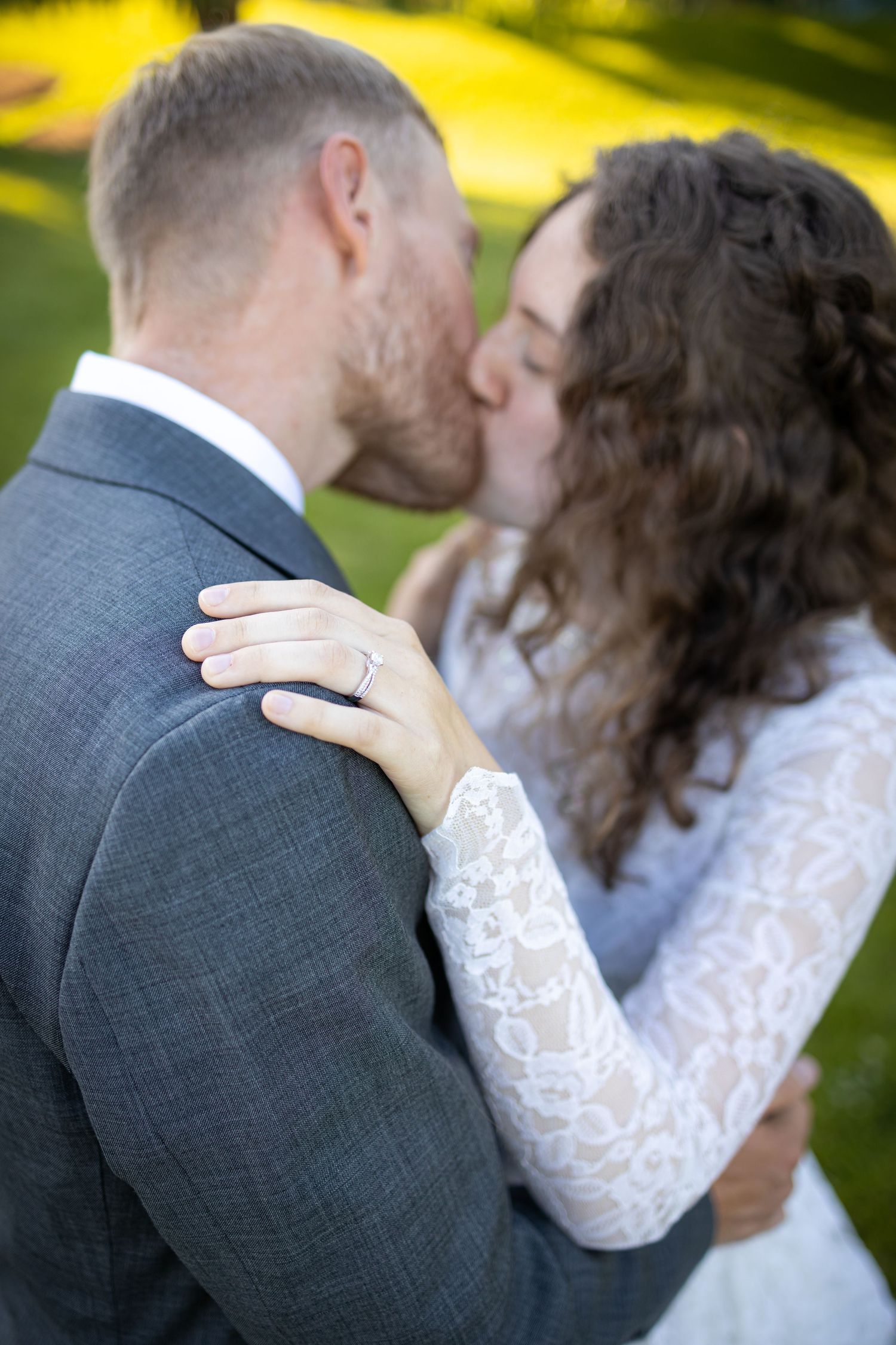 A romantic kiss between newlyweds outdoors with the bride's hand showing her ring on the groom's shoulder.