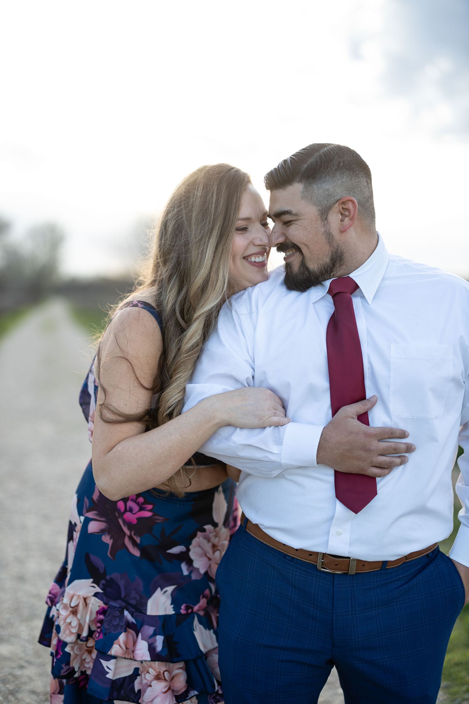 A romantic couple embraces on a path at sunset, she in a floral dress and he in dress shirt and red tie.