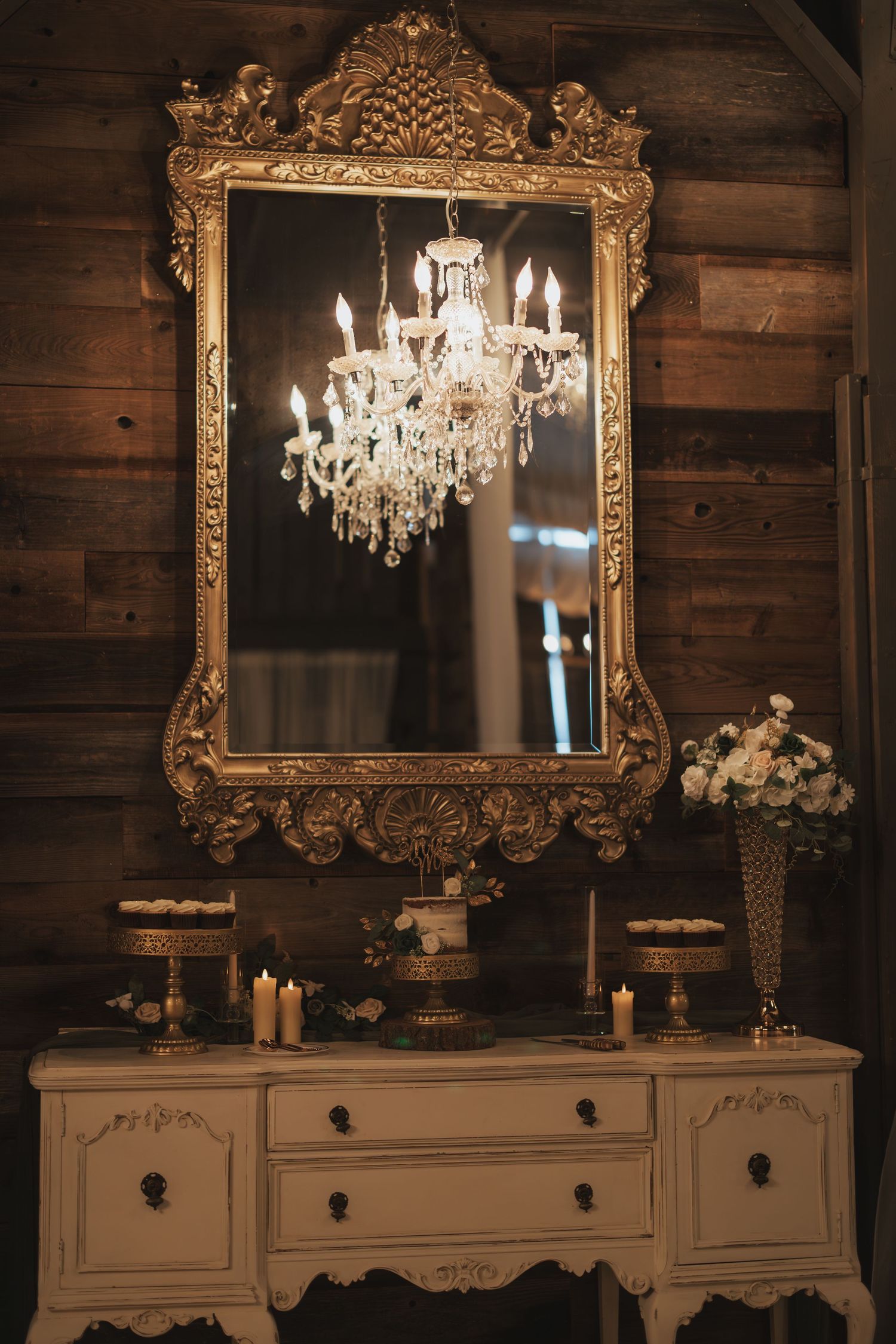 Ornate gold mirror reflects crystal chandelier against rustic wooden wall with vintage vanity below.