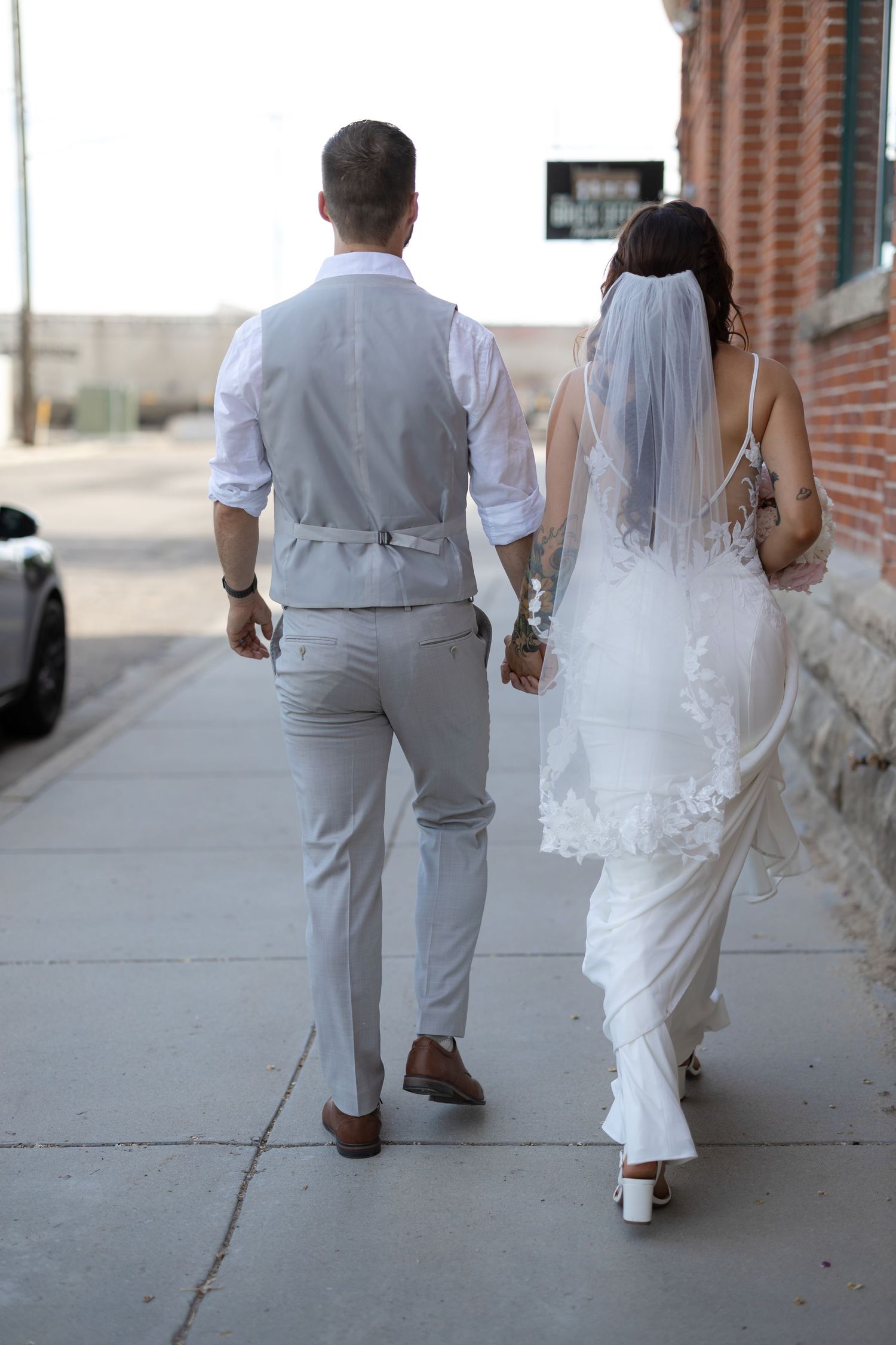 Bride and groom walking hand in hand down urban sidewalk showing backs of wedding attire.
