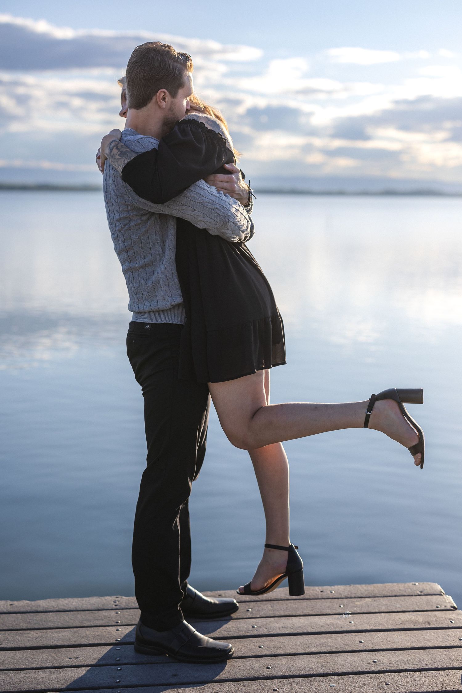 Couple sharing an intimate embrace on a wooden pier with lake and sky backdrop.