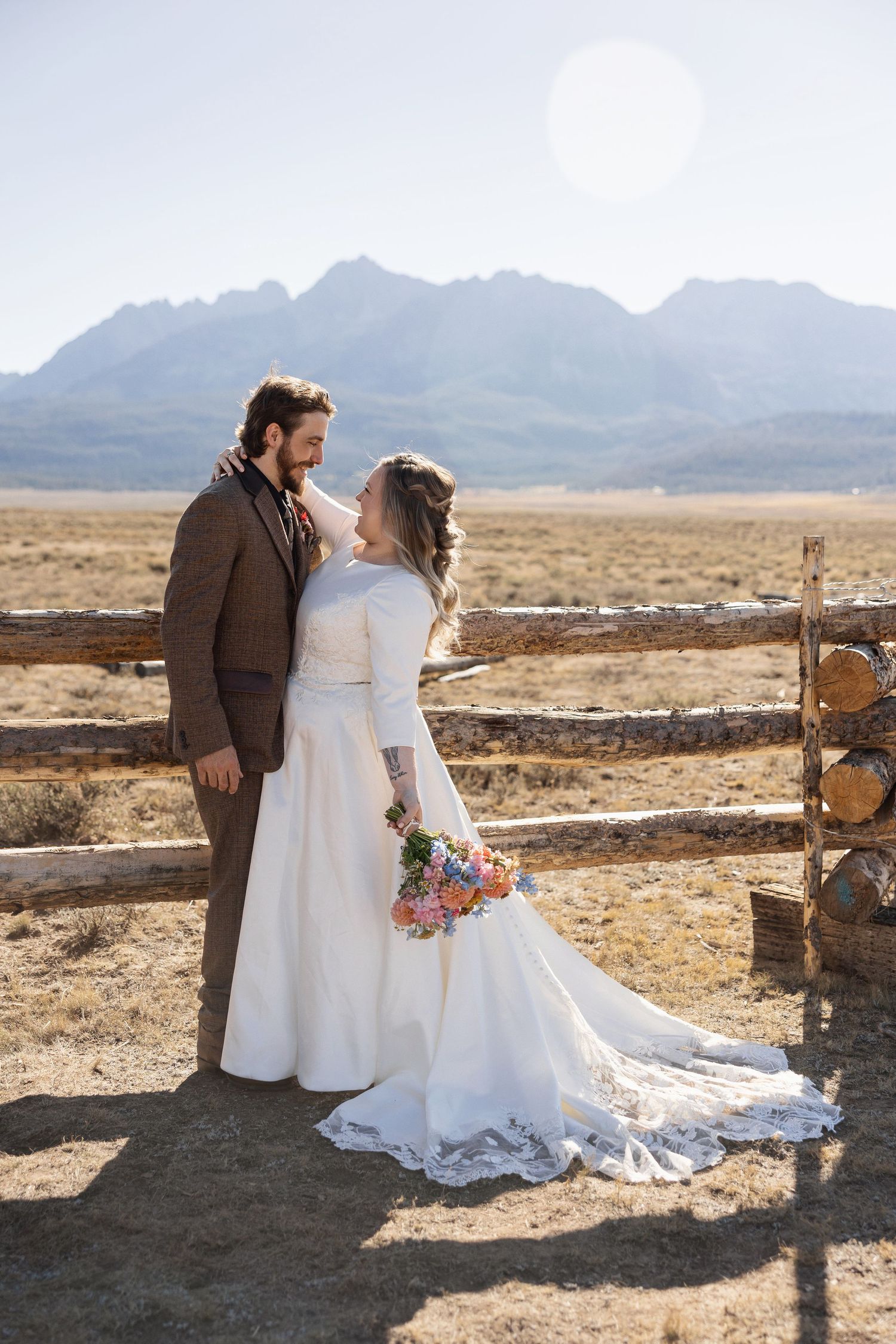 Newlyweds pose together against rustic wooden fence with mountain landscape in background.