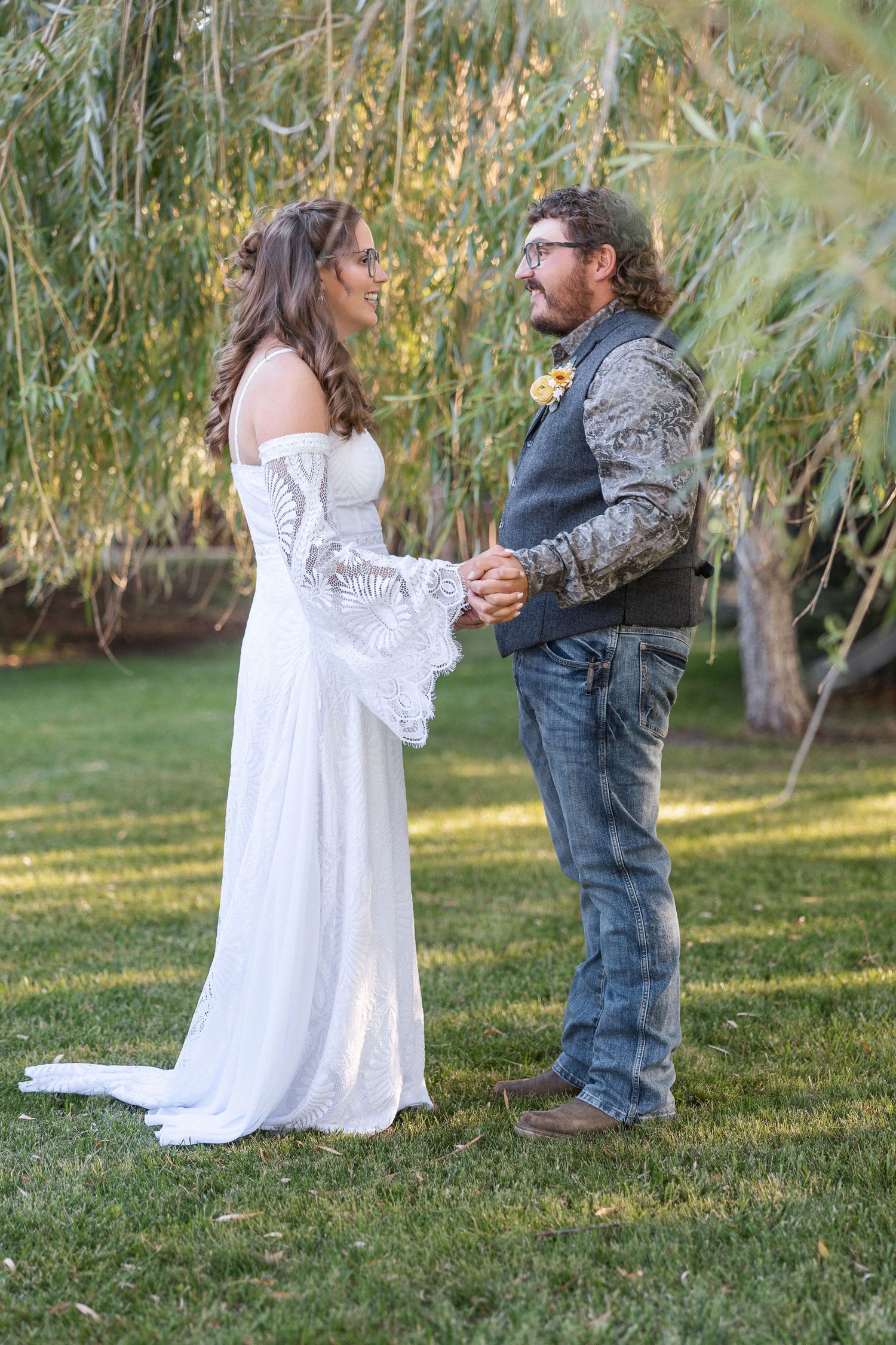 A romantic scene unfolds beneath willow branches as a couple in formal attire stands on green grass.