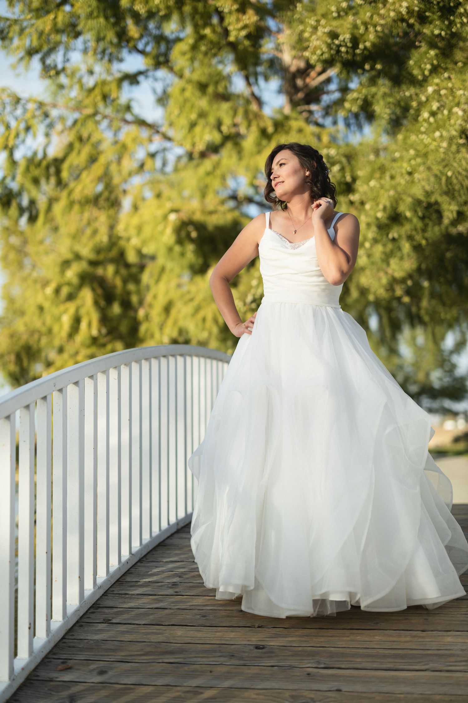Bride in flowing white gown poses elegantly on a wooden bridge with sunlit trees in background.
