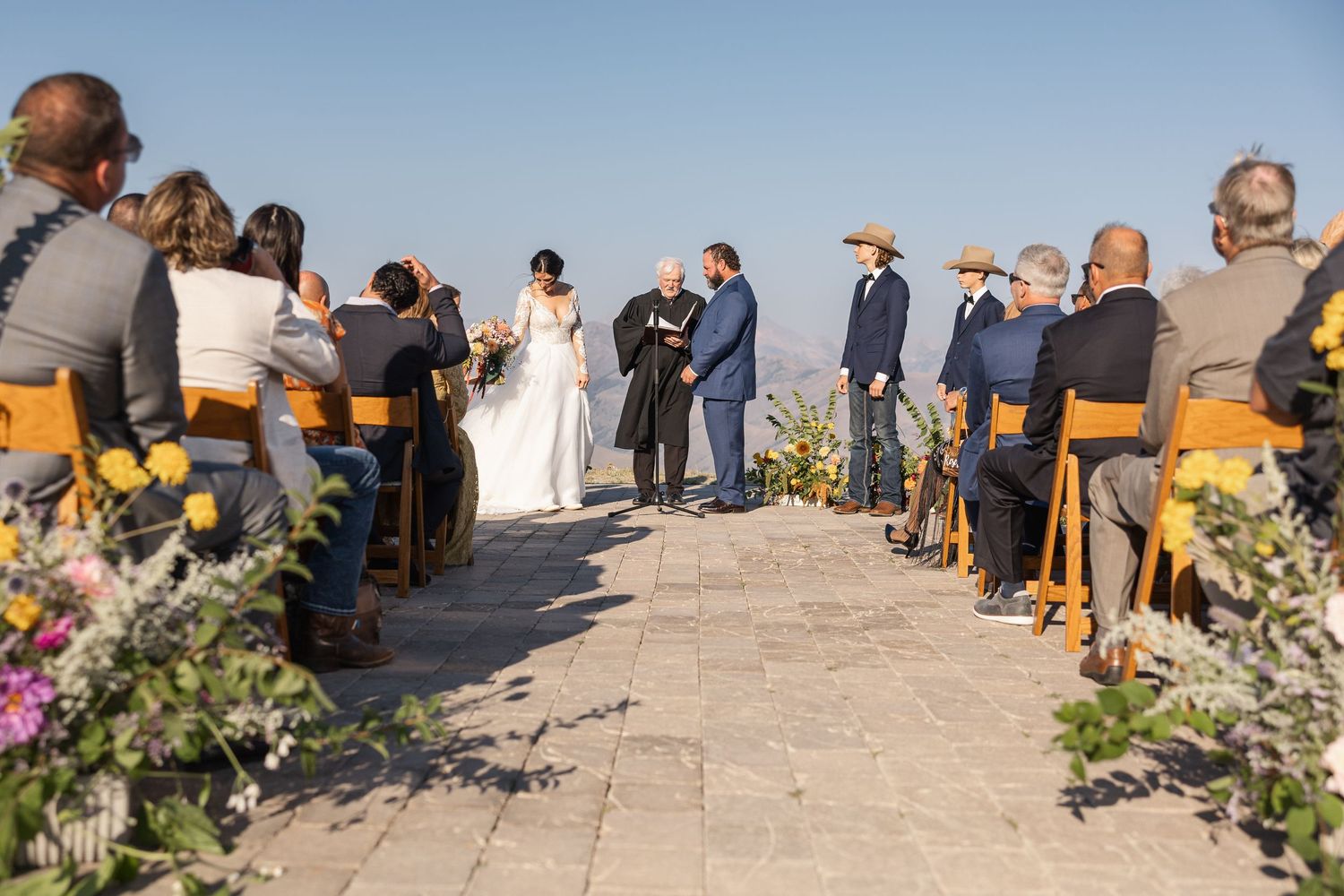A wedding ceremony aisle decorated with floral arrangements as guests watch the proceedings outdoors.