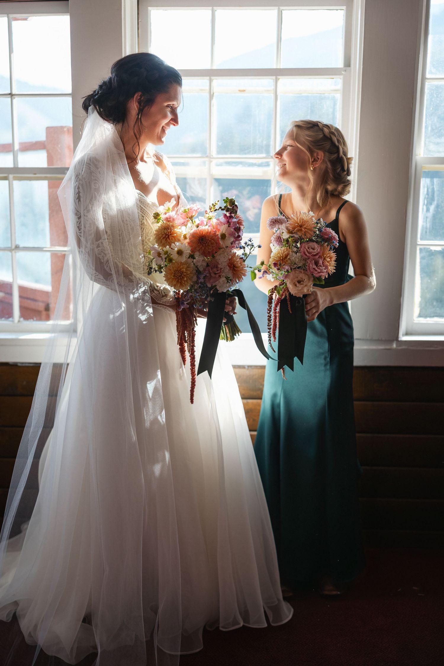 Wedding party members in white gowns and teal dresses holding floral bouquets by sunlit windows.