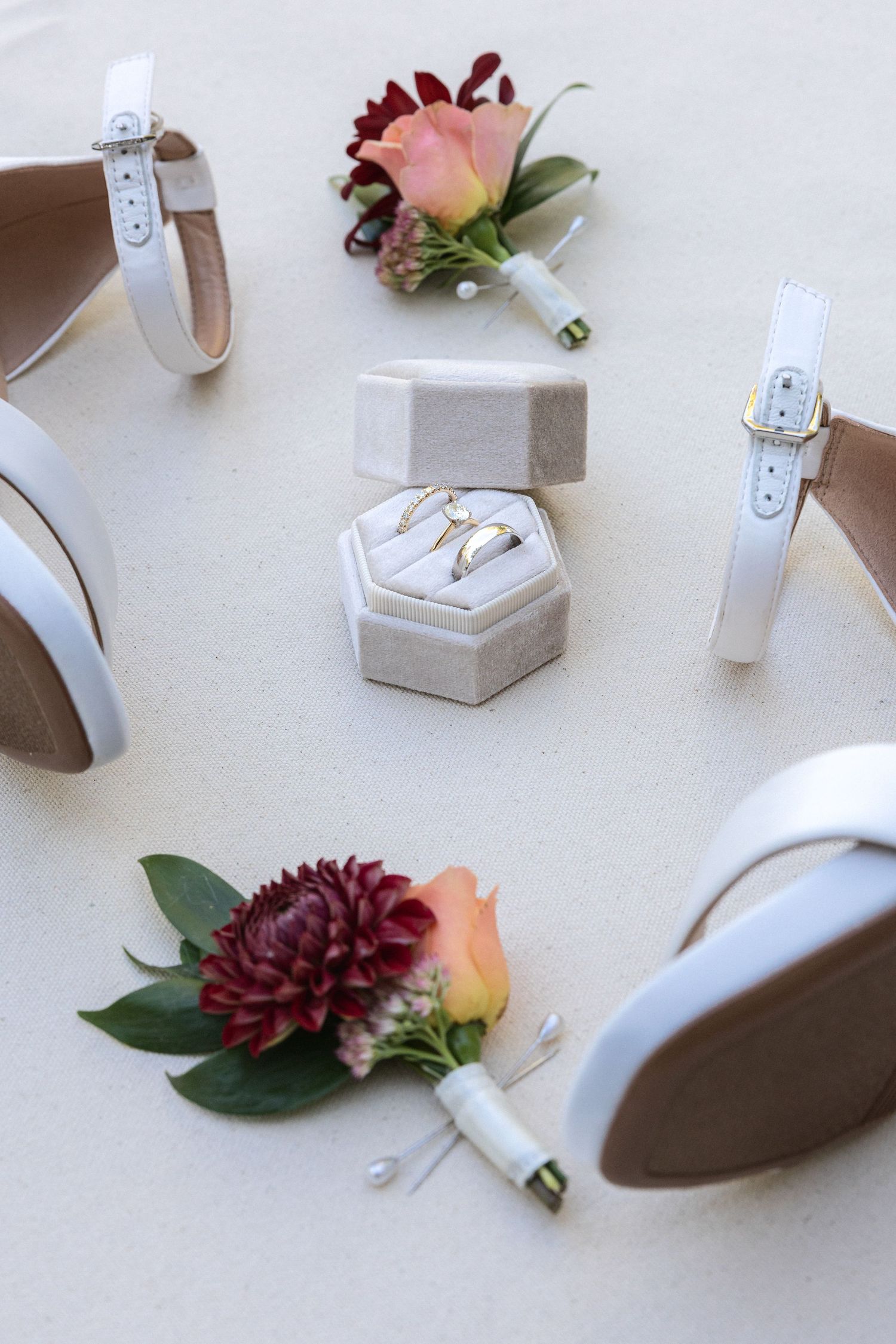Wedding details including rings, shoes, and burgundy florals arranged on white surface.
